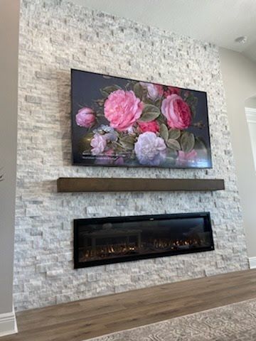 Large TV displaying floral art, above a fireplace with a wooden beam, mounted on a white stone wall.