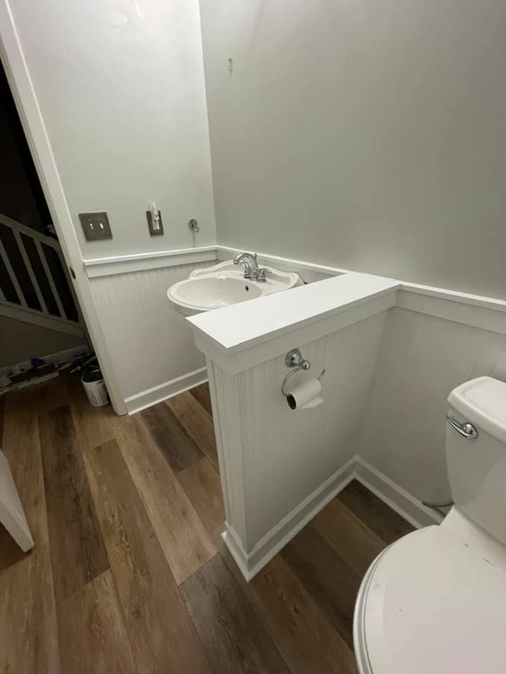 A small, white bathroom with a sink, toilet, and wood-look flooring. The sink is on a half-wall.