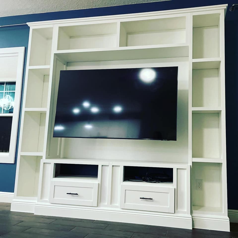 White built-in entertainment center with TV, shelves, drawers, against a dark blue wall.