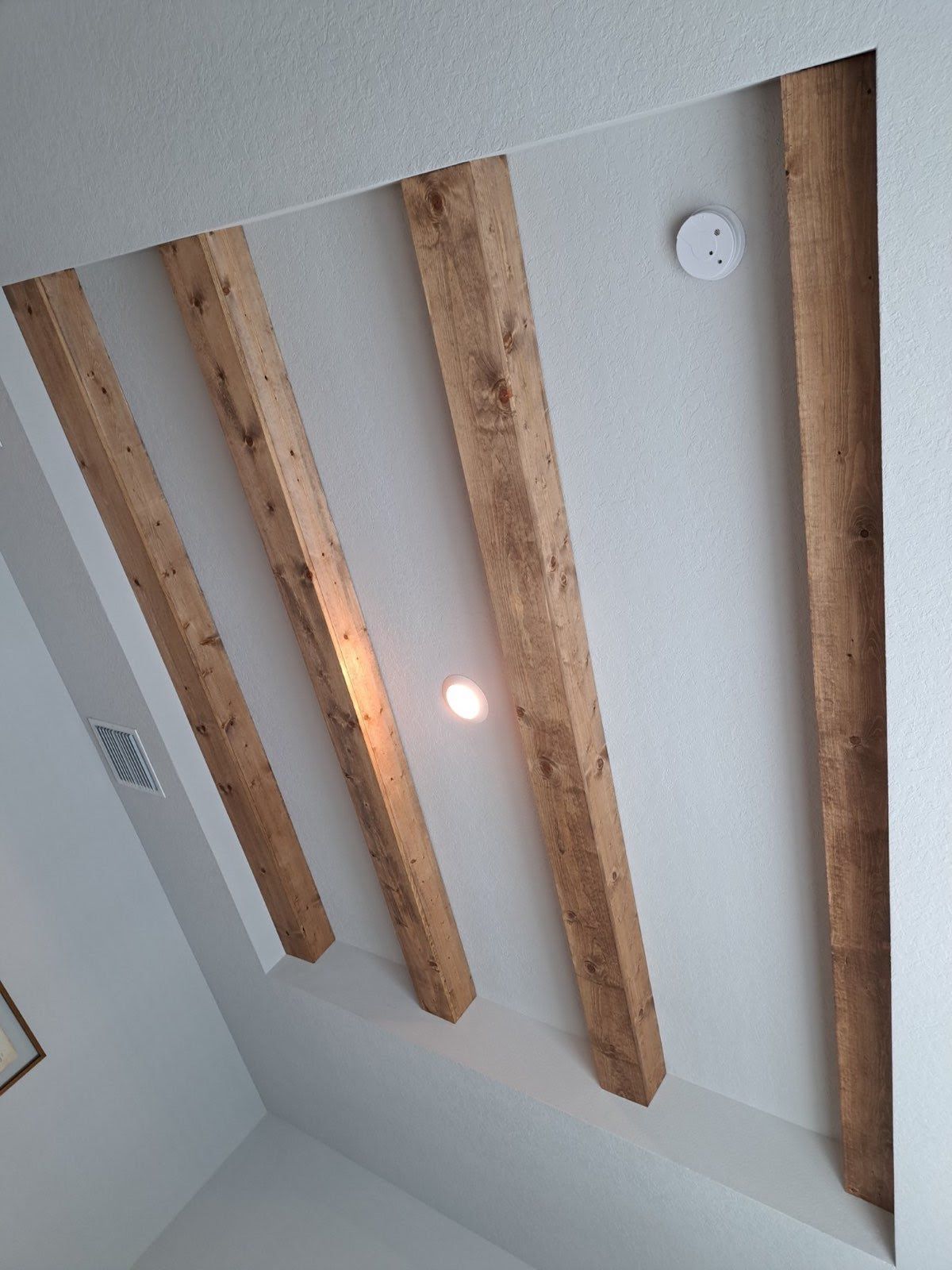 Wooden beams on a white ceiling with a light fixture and smoke detector.
