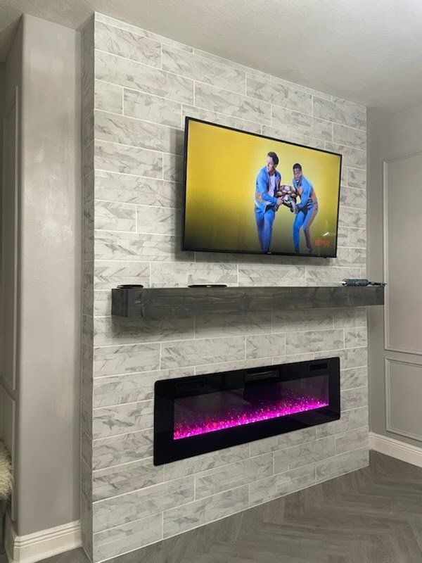 A TV, fireplace, and wooden shelf on a gray brick wall; a couple is on the TV screen.