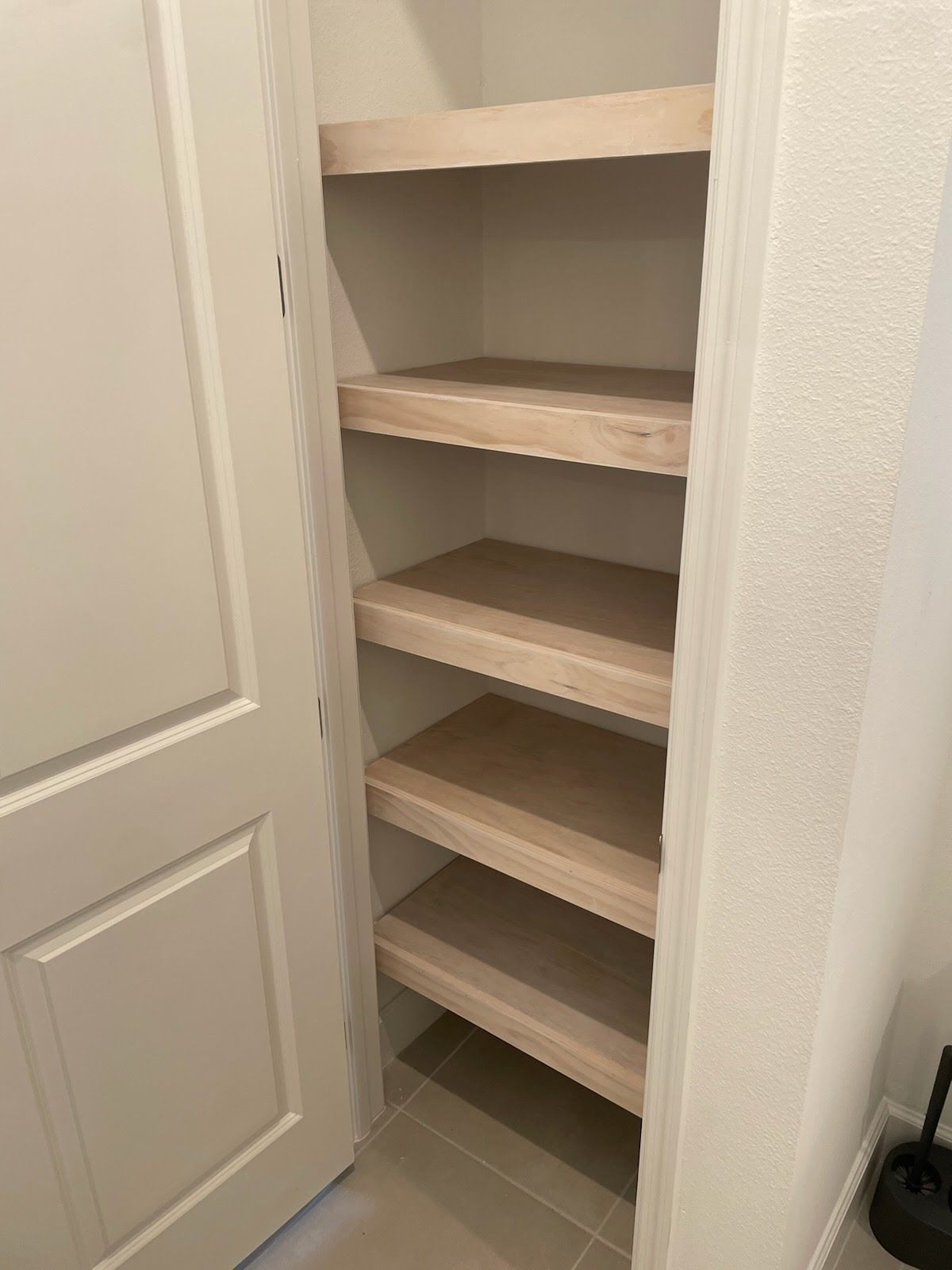 Empty built-in pantry with five wooden shelves, light wood tone, inside a white-walled closet, beige tile floor.