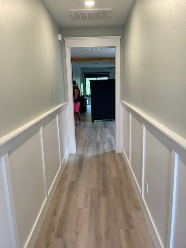 Hallway with gray-blue walls, white wainscoting, light wood flooring, and doorway to a brighter room.