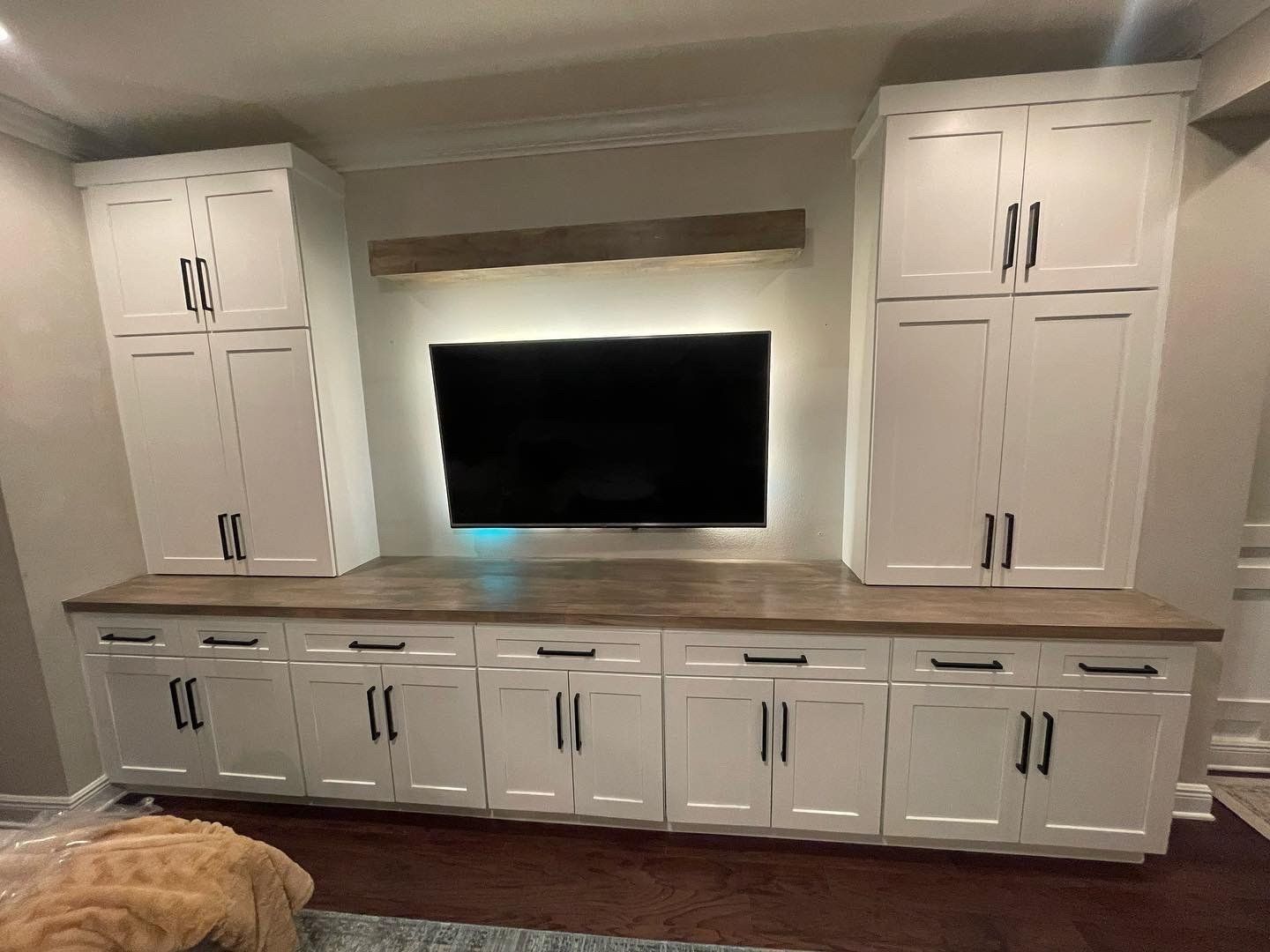 White built-in entertainment center with TV, cabinets, and dark hardware, in a room with dark flooring.
