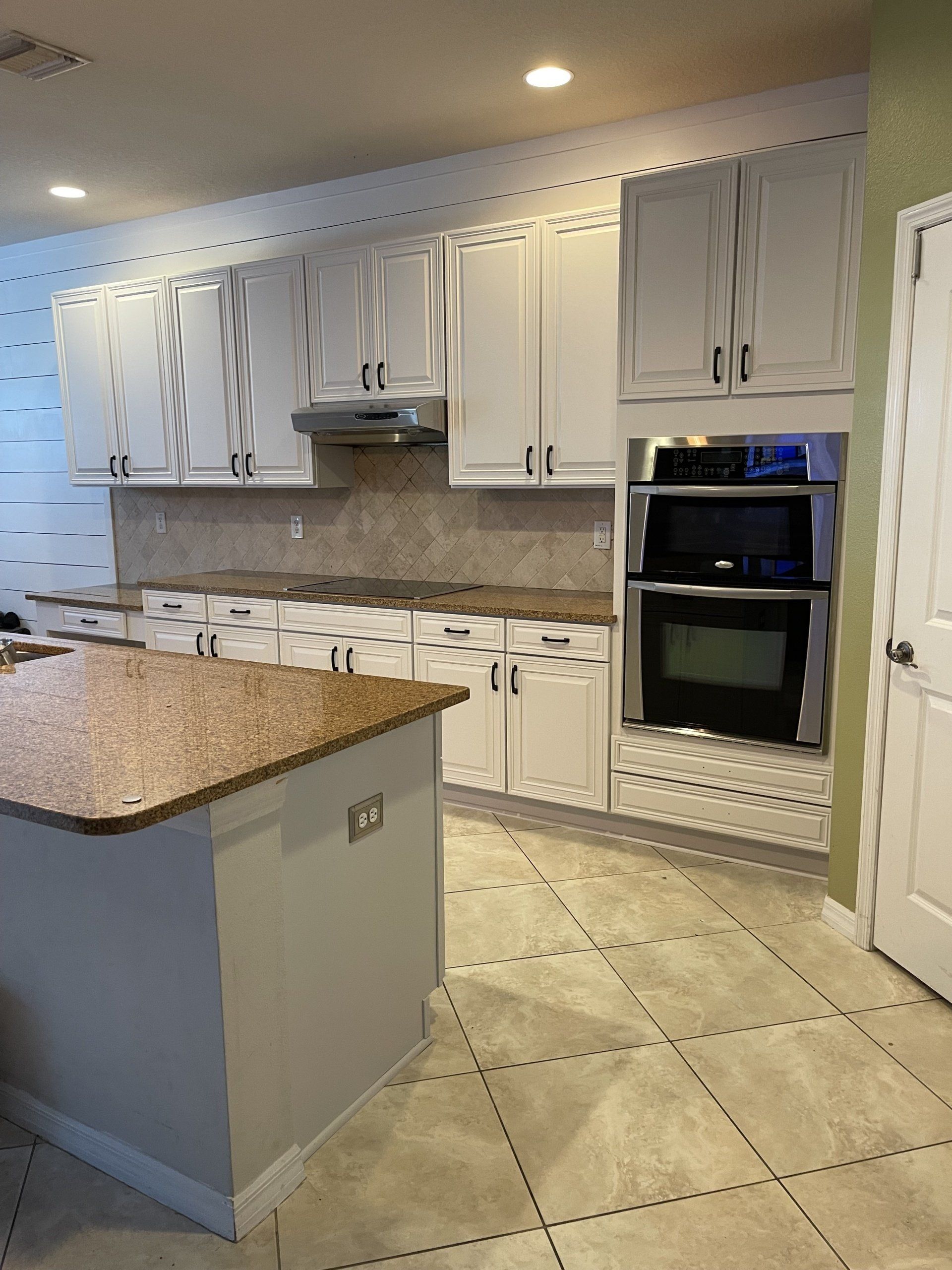 White kitchen cabinets and island with granite countertops, double oven, and beige tiled floor.