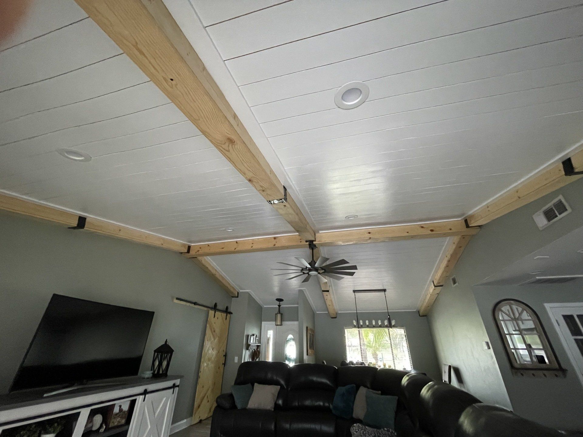 Living room ceiling with white paneling and wood beams, a TV, and a dark-colored sectional sofa.