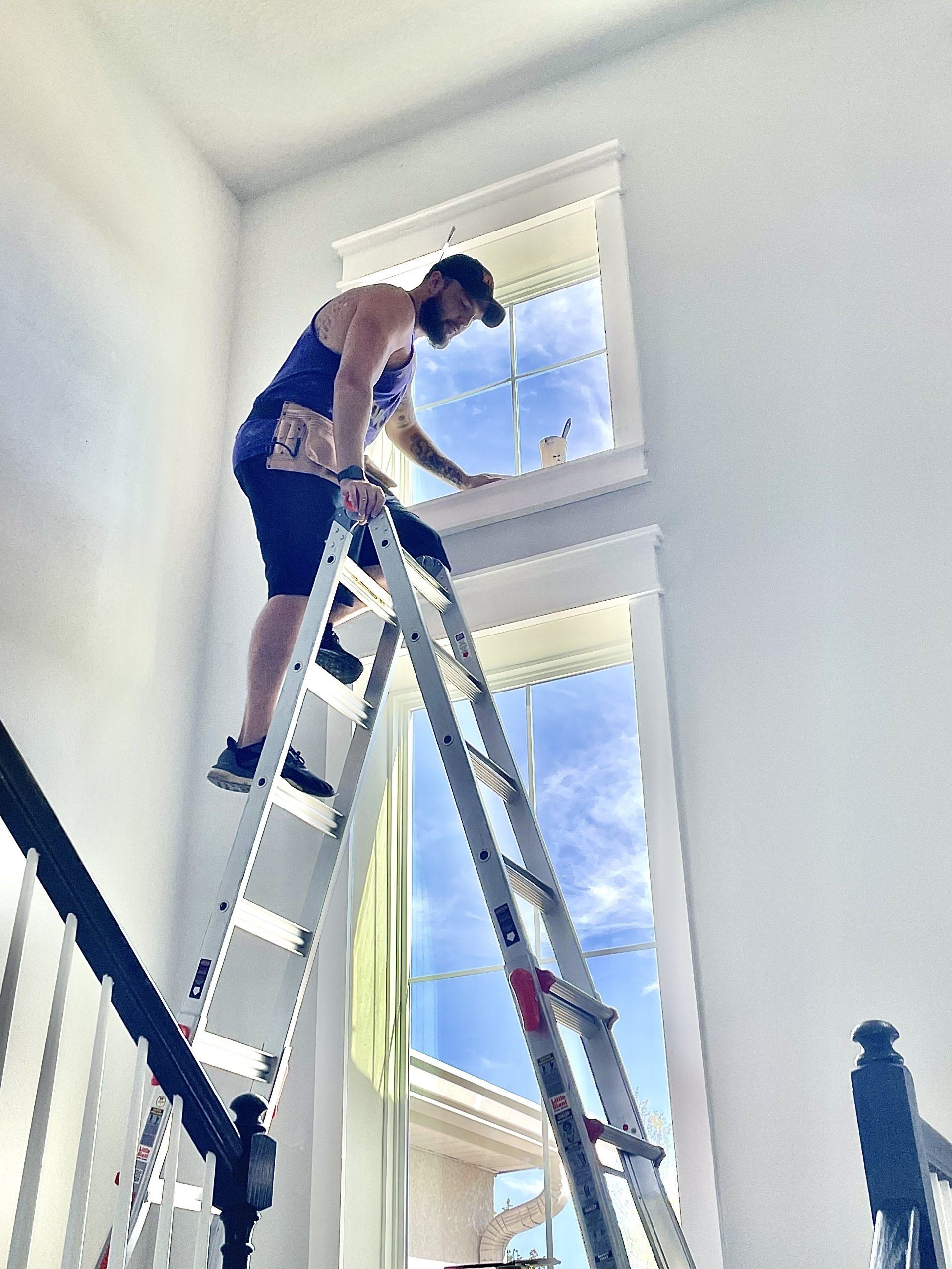 Man on a ladder cleaning a tall window with a blue sky in the background.