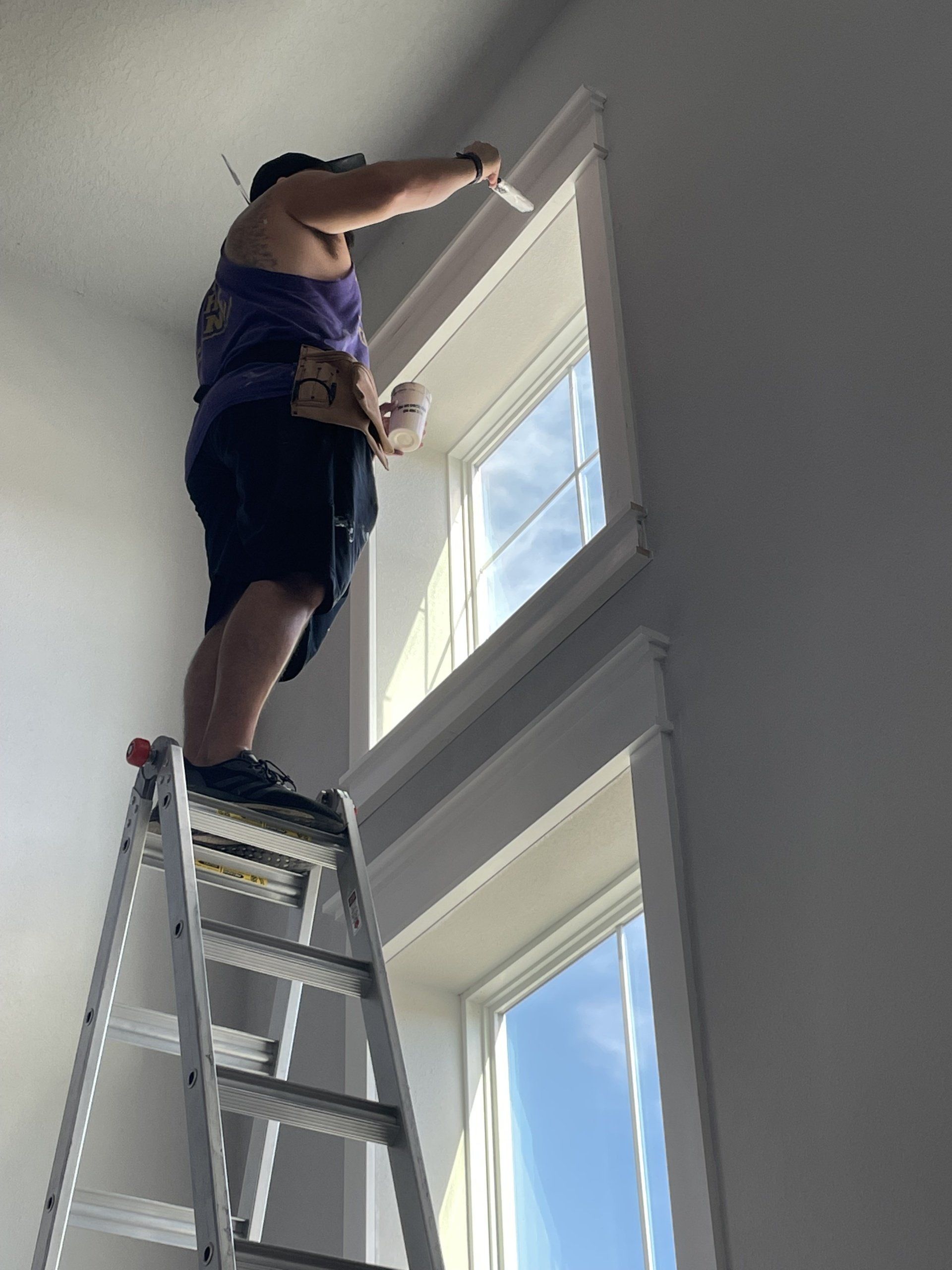 Man on a ladder installing trim around a tall window.