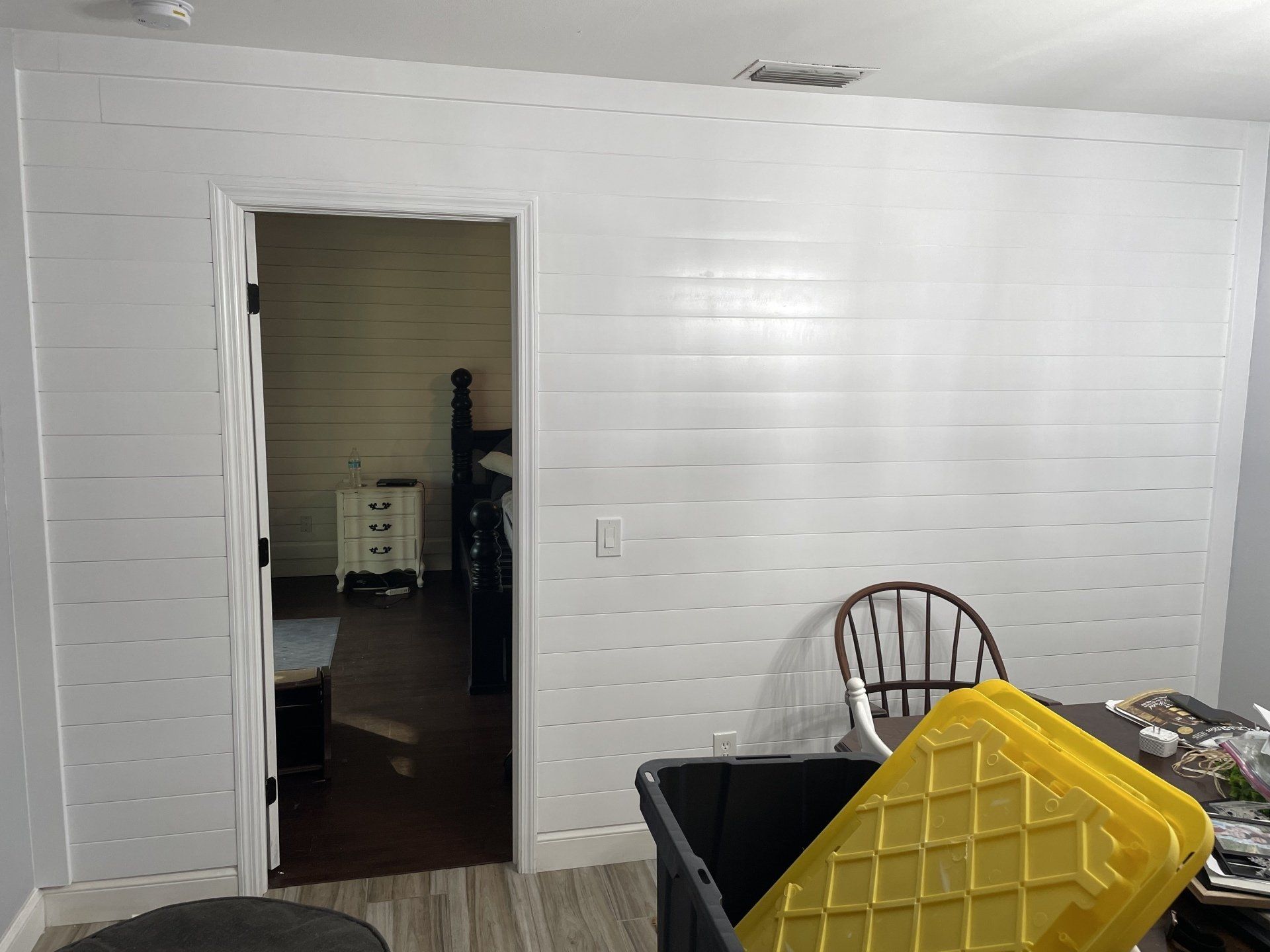 White paneled wall with an open door to a bedroom. A yellow bin sits in front of the wall, and a chair.