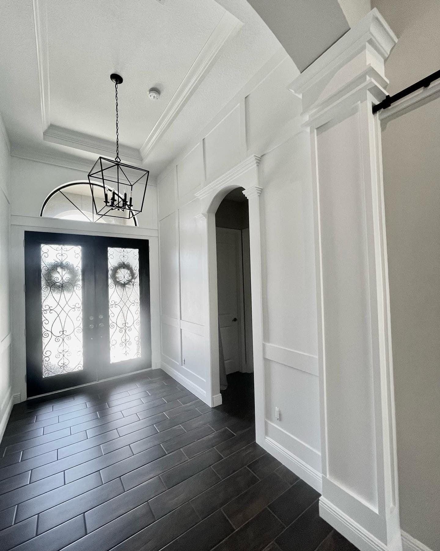 Elegant white entryway with dark wood floor and chandelier. Black door, arched doorway, wainscoting.