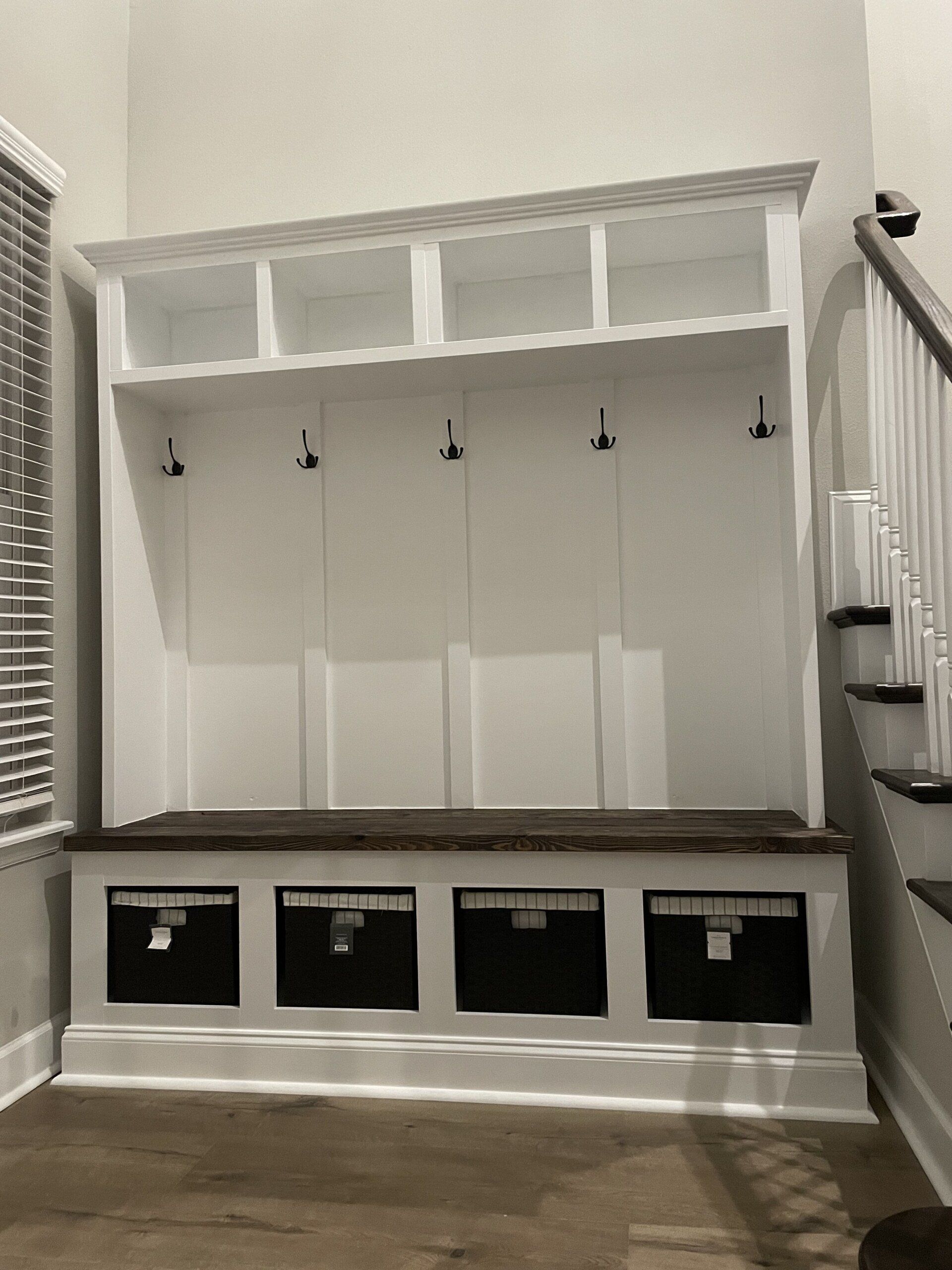 White built-in entryway storage unit with bench, cubbies, hooks, and black baskets against a wall.