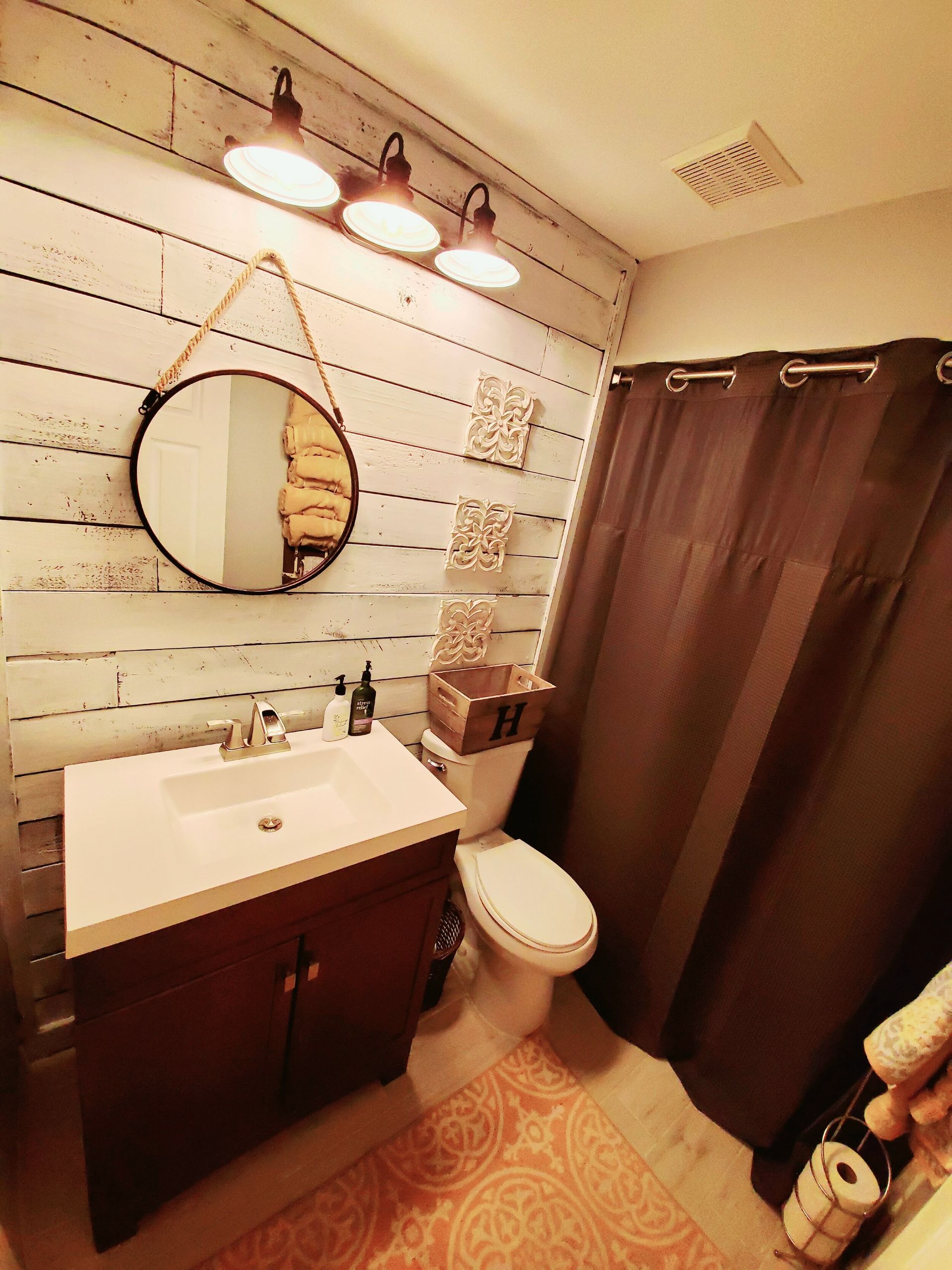 Bathroom with wood-paneled wall, round mirror, brown vanity, toilet, shower curtain, and rustic light fixture.