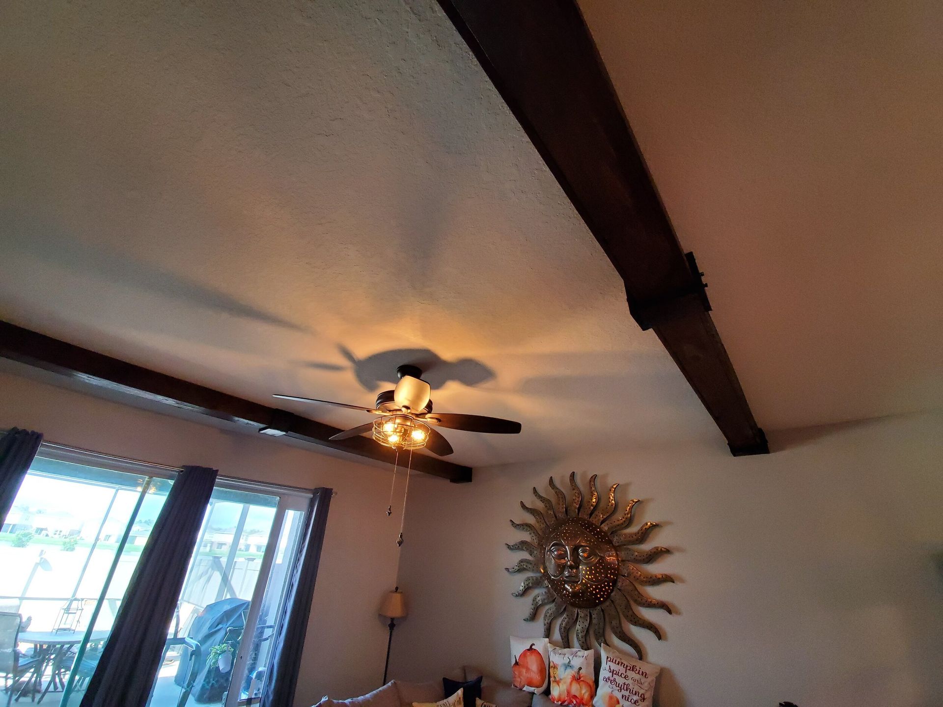 Living room ceiling with dark beams, a ceiling fan, and sunburst wall decor.