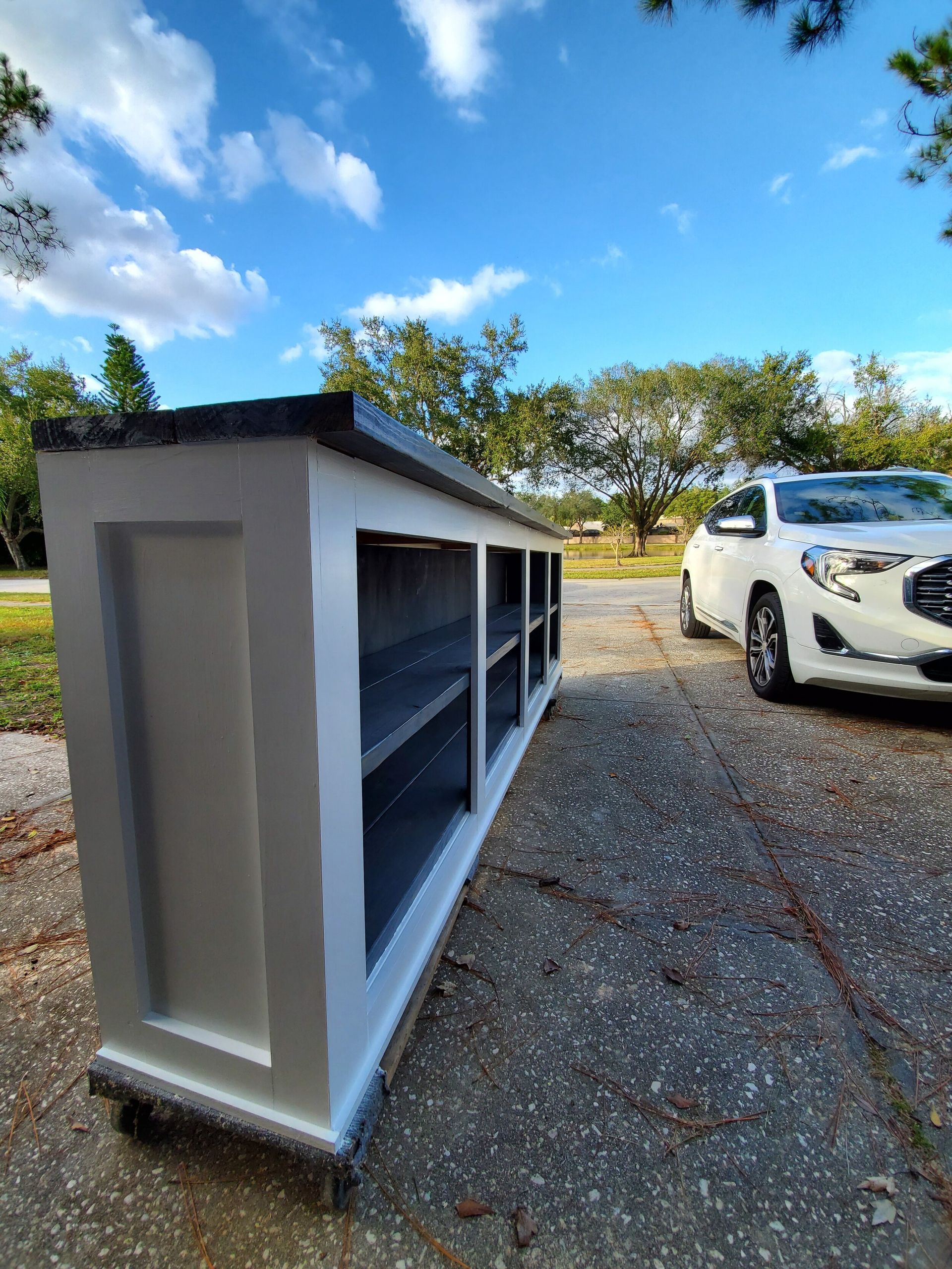 White cabinet with gray interior and countertop, outdoors on a paved surface, with a car and trees in the background.