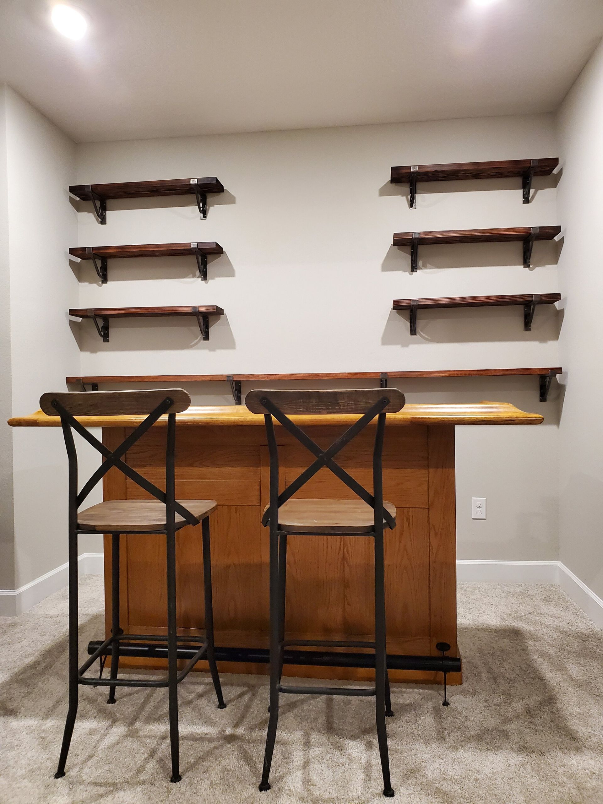 Wooden bar with two stools and shelves against a beige wall in a room with carpet.