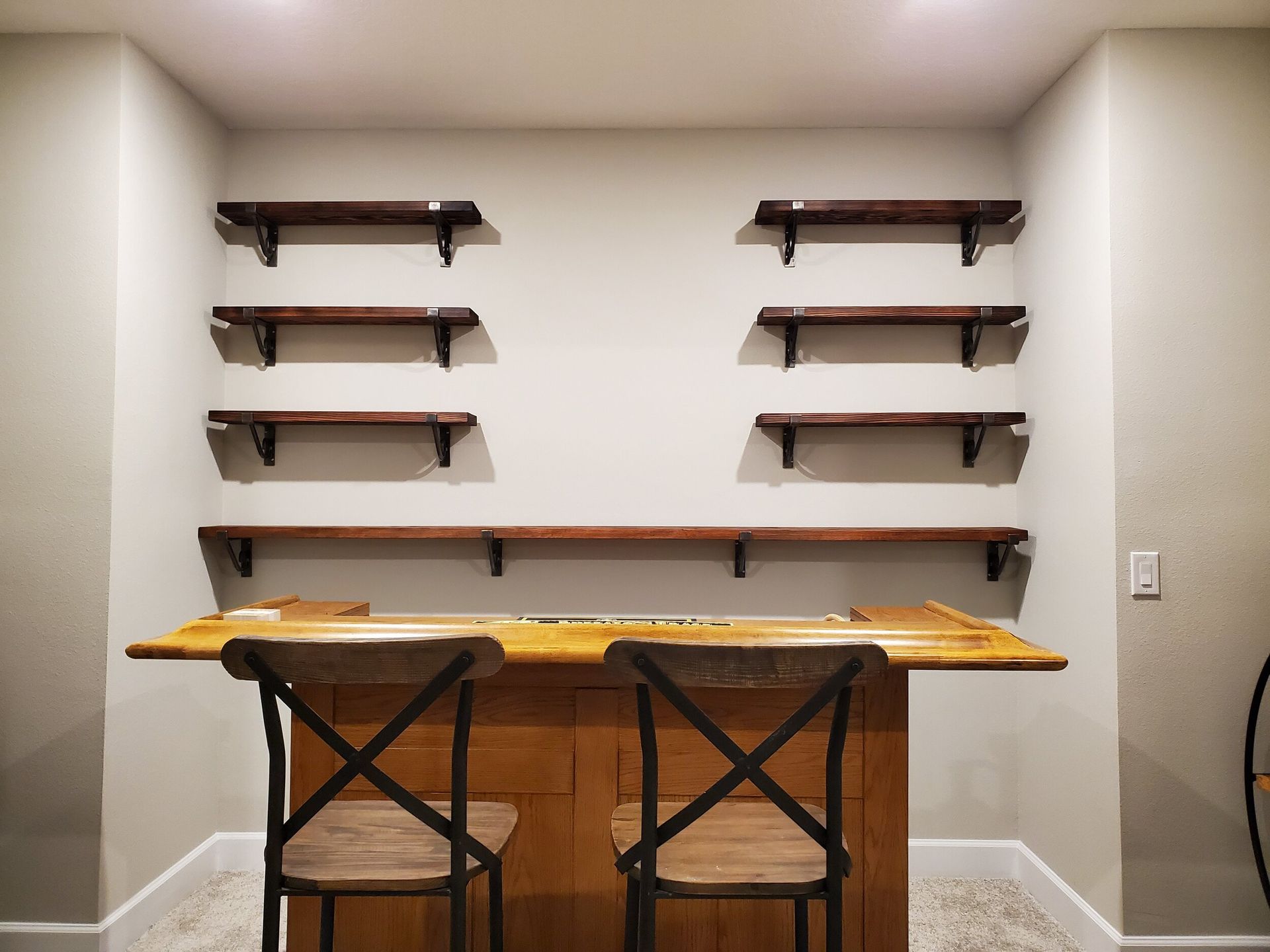 A bar with shelves, two chairs, and a light gray wall. The bar is brown, and so are the shelves.