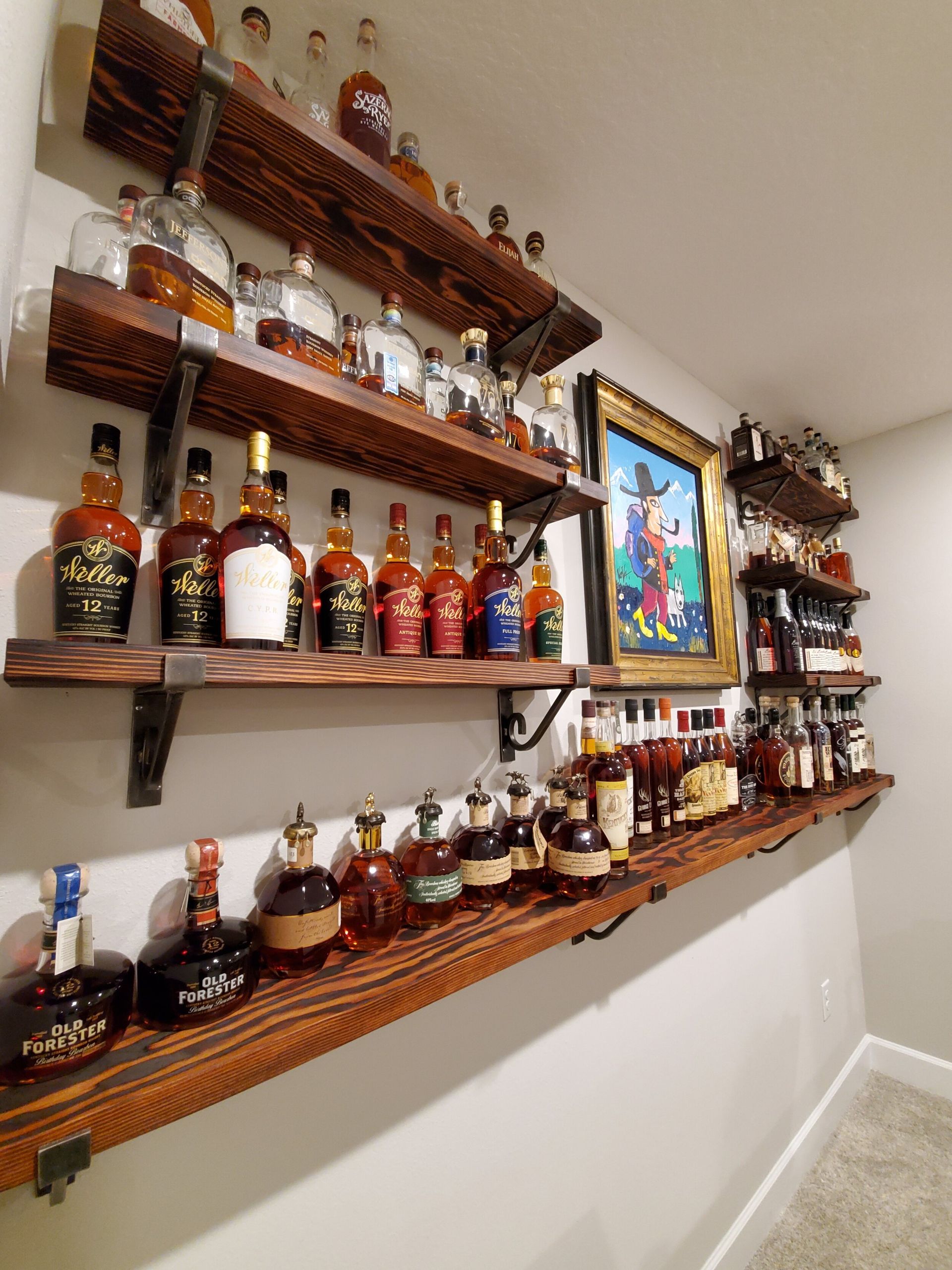 Wooden shelves displaying various bourbon bottles on a light gray wall. Artwork hangs between the shelves.