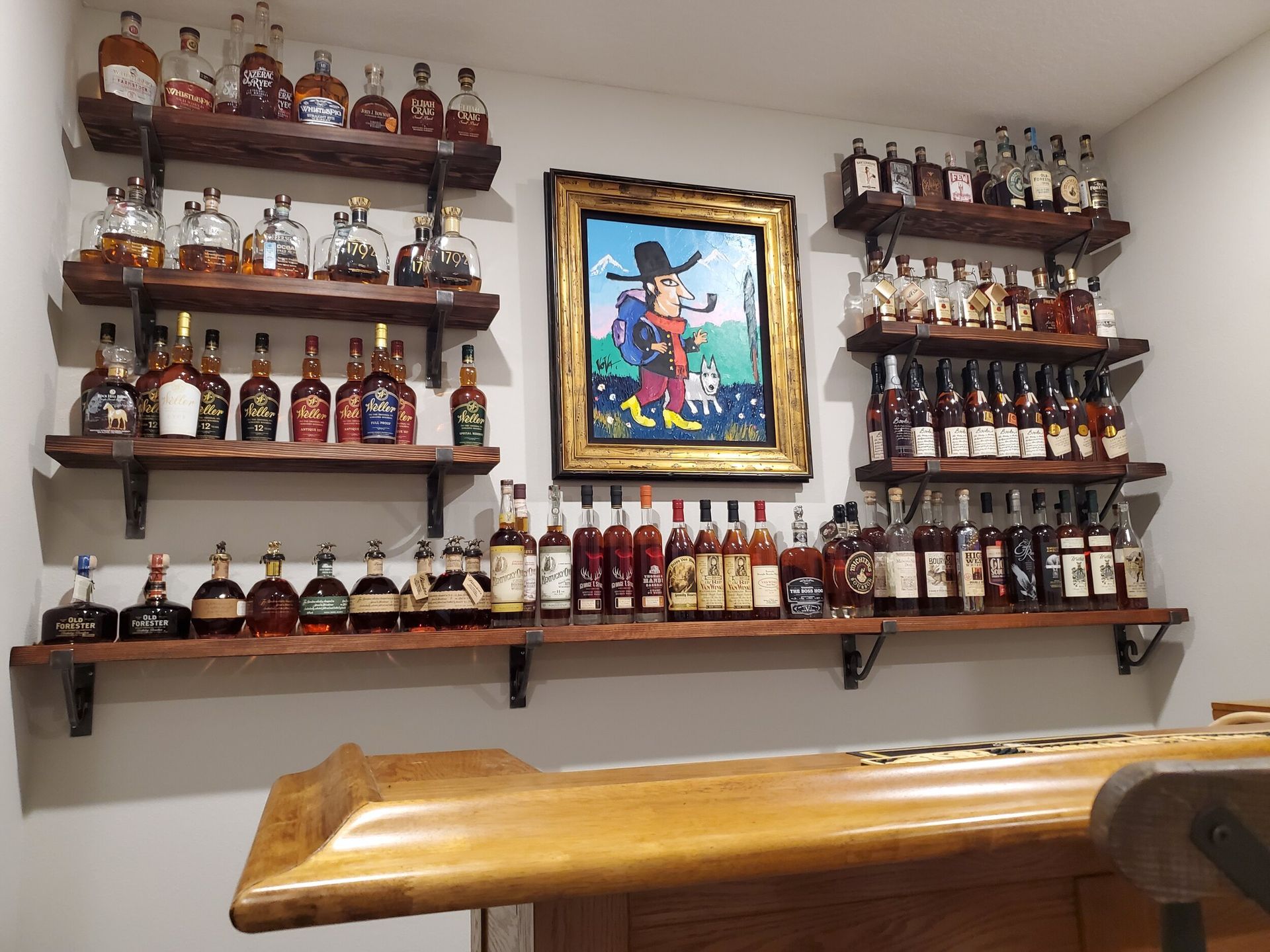 Wooden shelves displaying liquor bottles above a wood bar and a painting on a white wall.