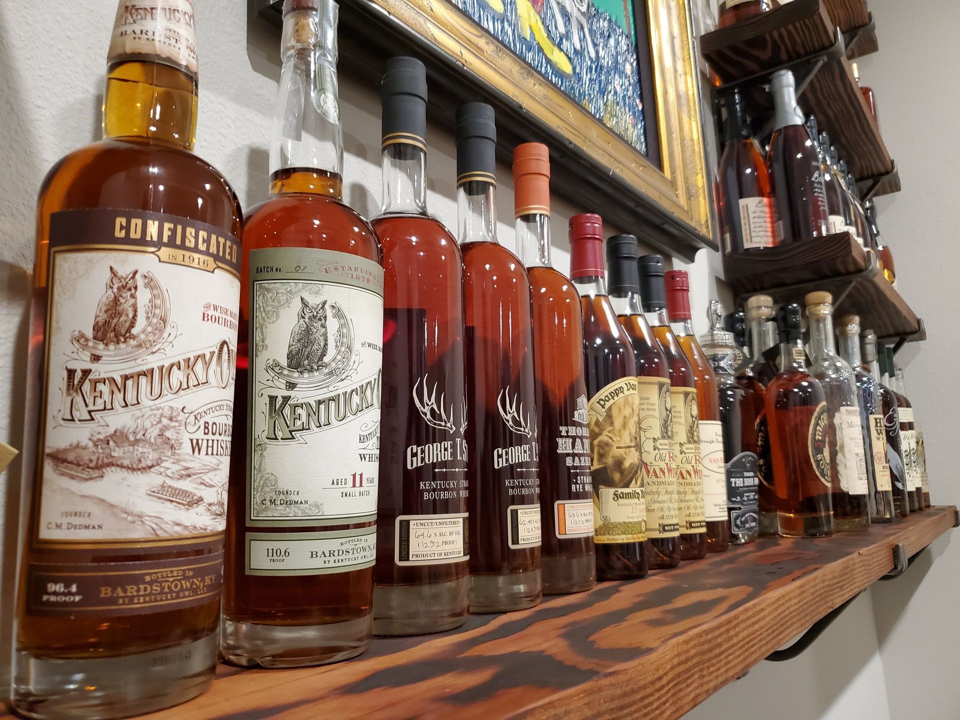 Whiskey bottles on a wooden shelf against a white wall, with a painting above.
