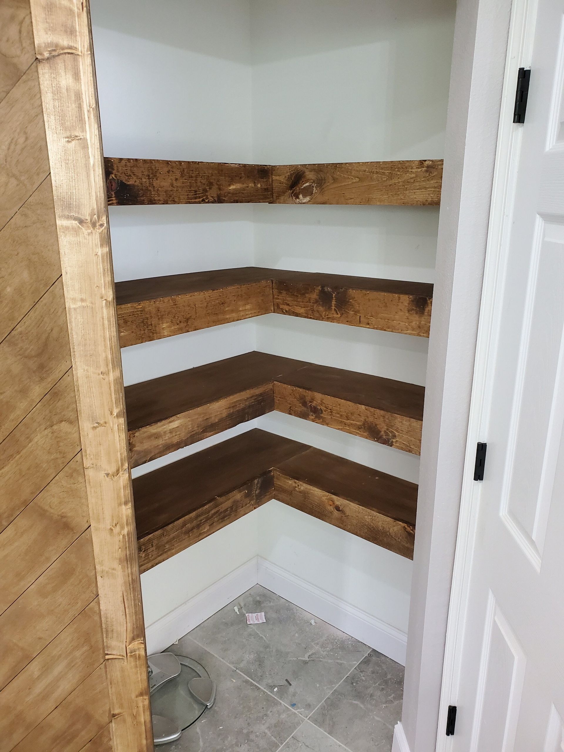 Corner pantry with three rustic, wooden shelves. White walls and door.