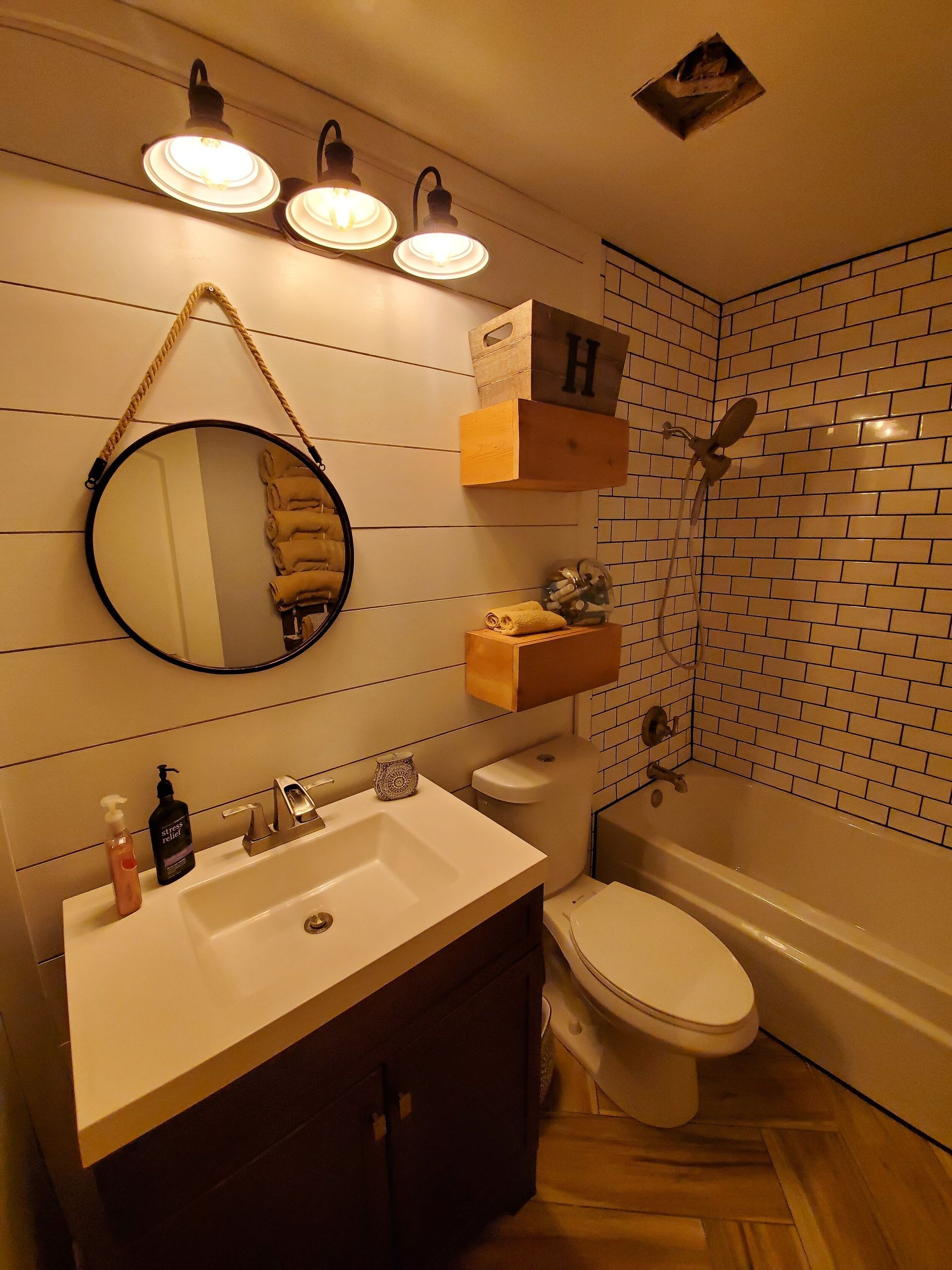 Bathroom with a white vanity, round mirror, and wooden accents. White tiled shower and wooden shelves.