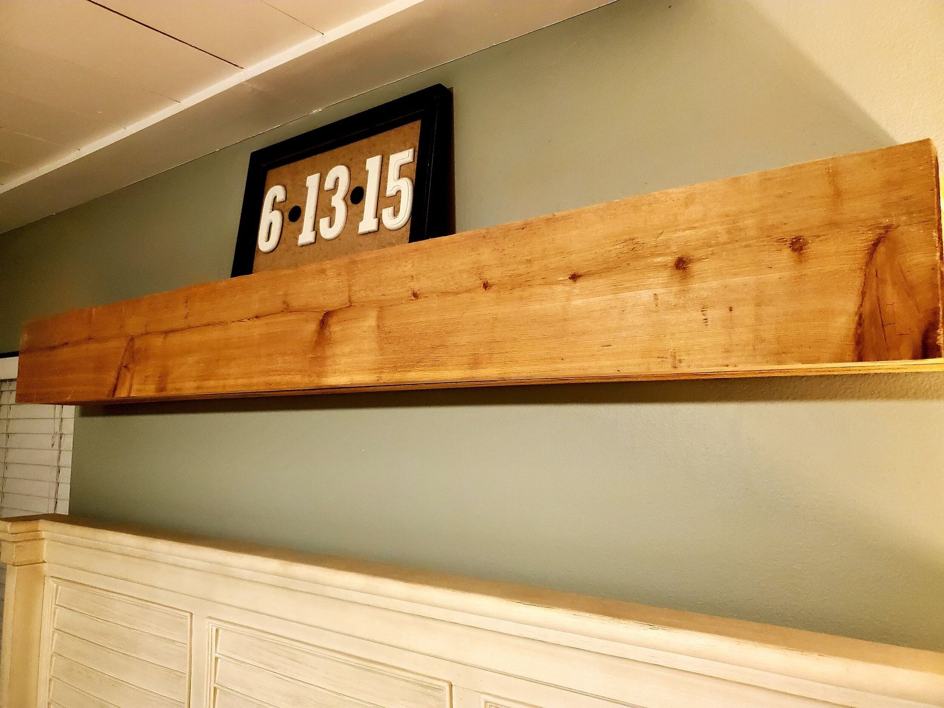 Wooden shelf mounted on a wall with a black framed sign above it; below is a cream-colored headboard.