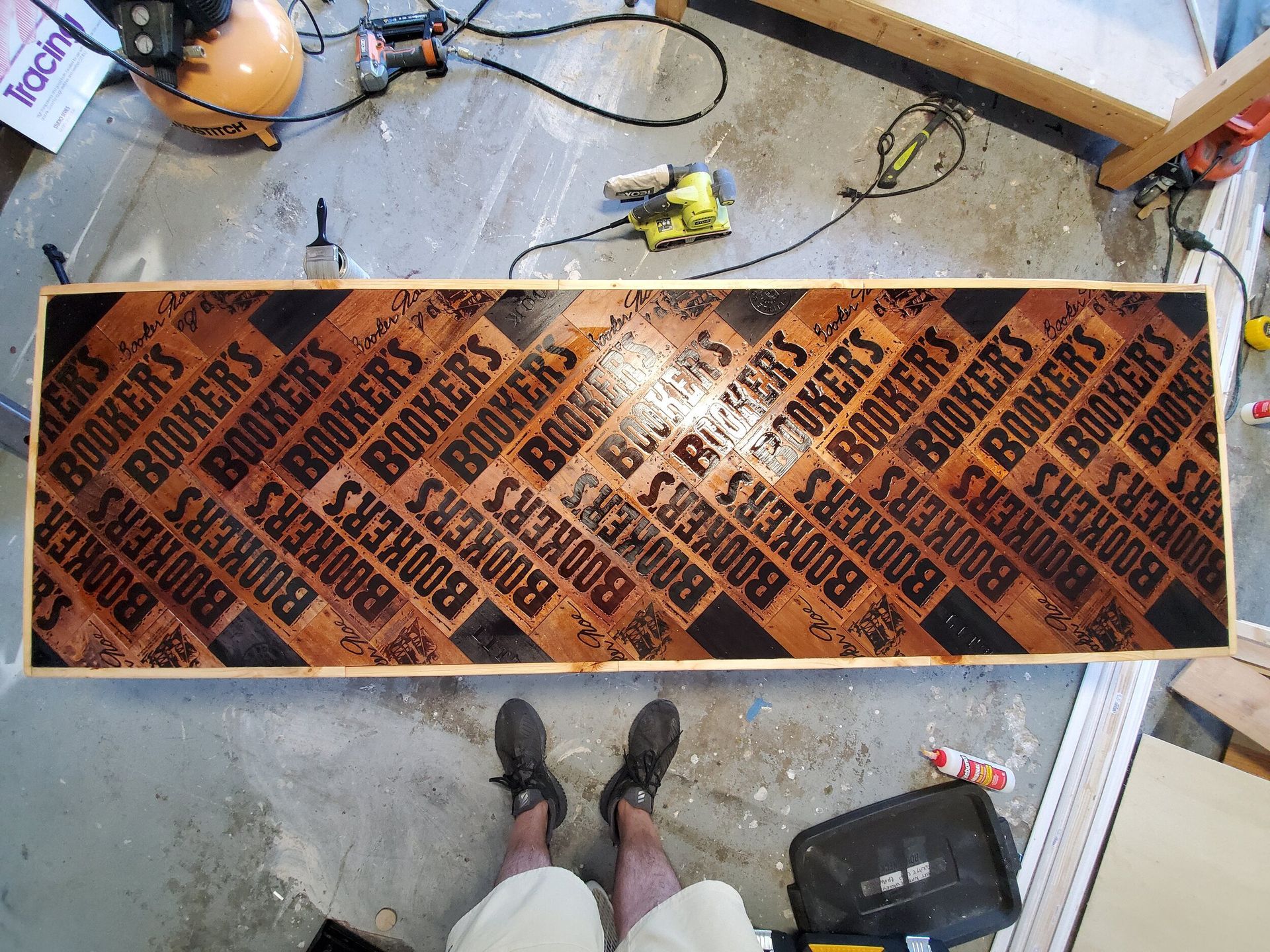 A wood table top with “BOOKER'S” branded designs. Tools and a person's feet are nearby.