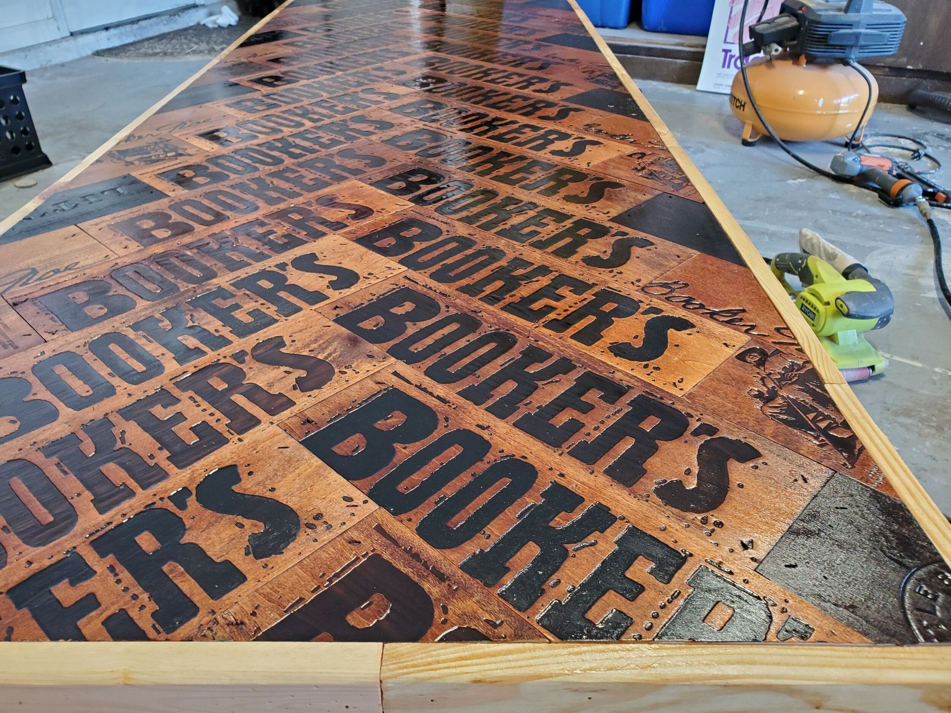 Booker's bourbon bottle label-themed bar top. Brown, black, and gold resin finish. Wooden frame, workshop setting.
