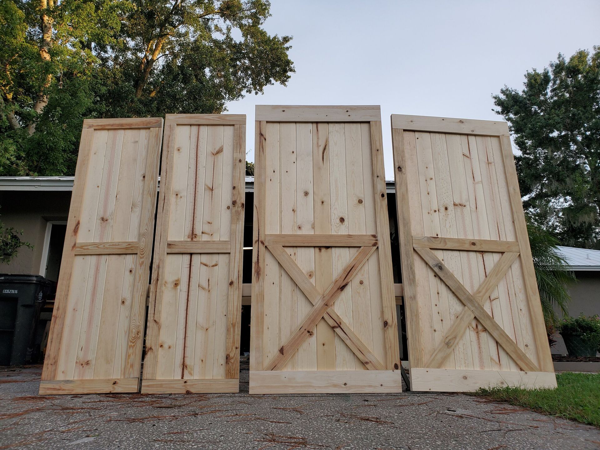 Four rustic, wooden barn doors leaning against a driveway in front of a house.