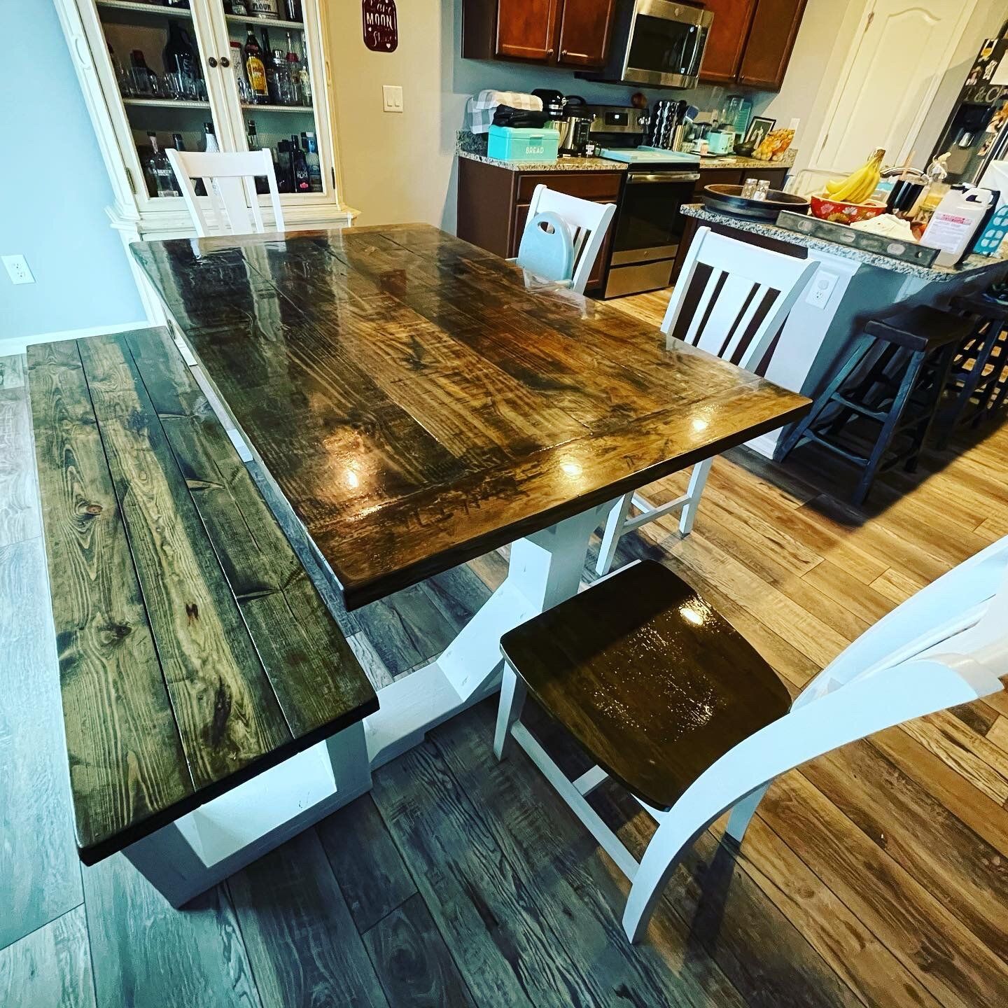 Rustic dining table and bench with white chairs in a kitchen with hardwood floors and a white cabinet.