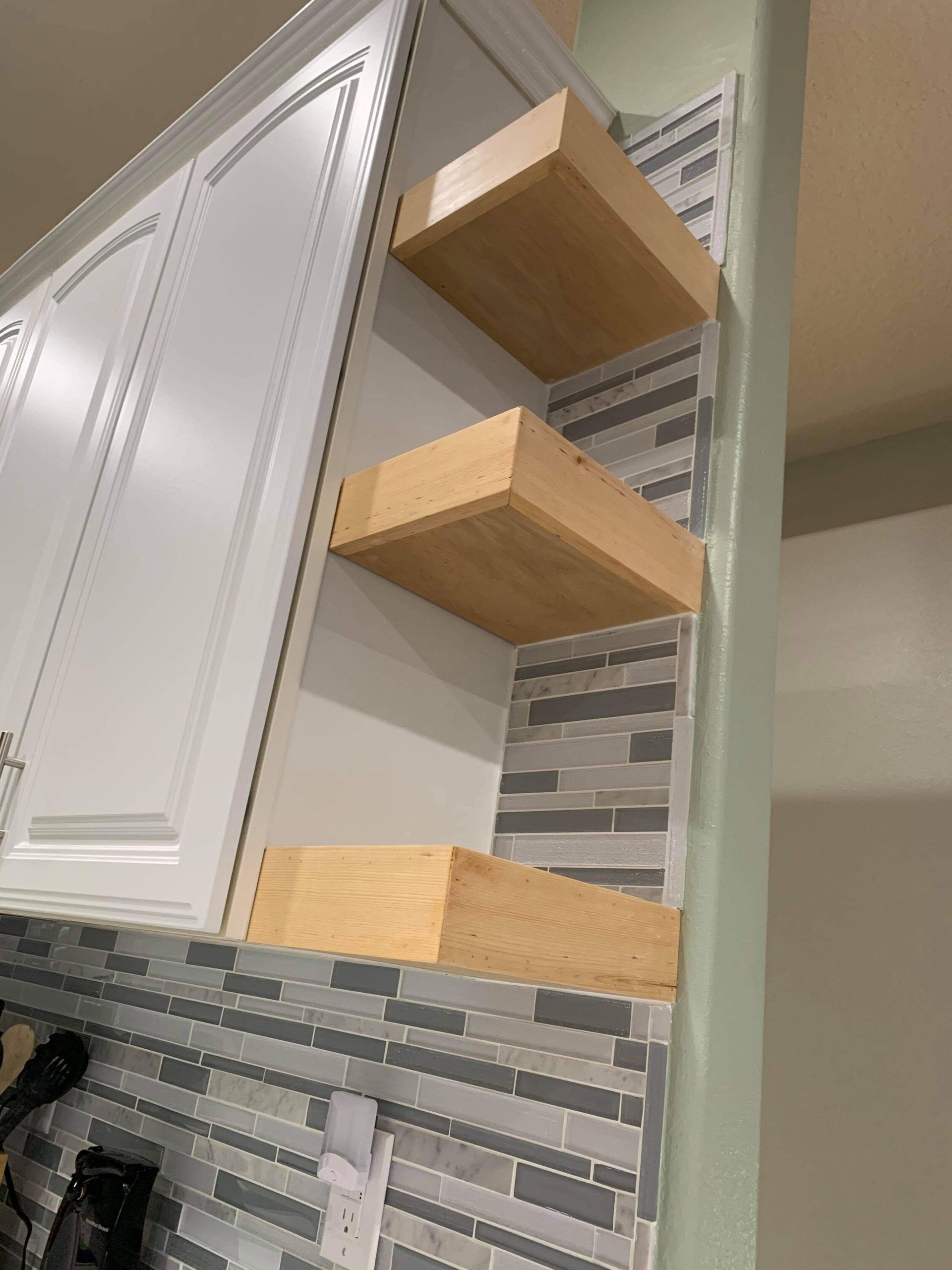 Three wooden shelves mounted on a tile backsplash next to a white cabinet and a green wall.