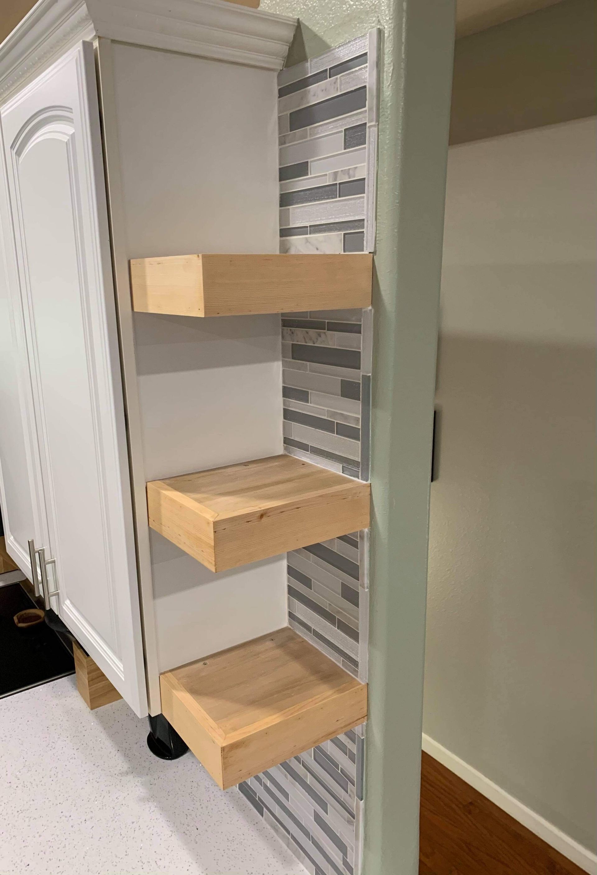 Three wooden shelves built into a kitchen cabinet, with a gray-tiled backsplash.