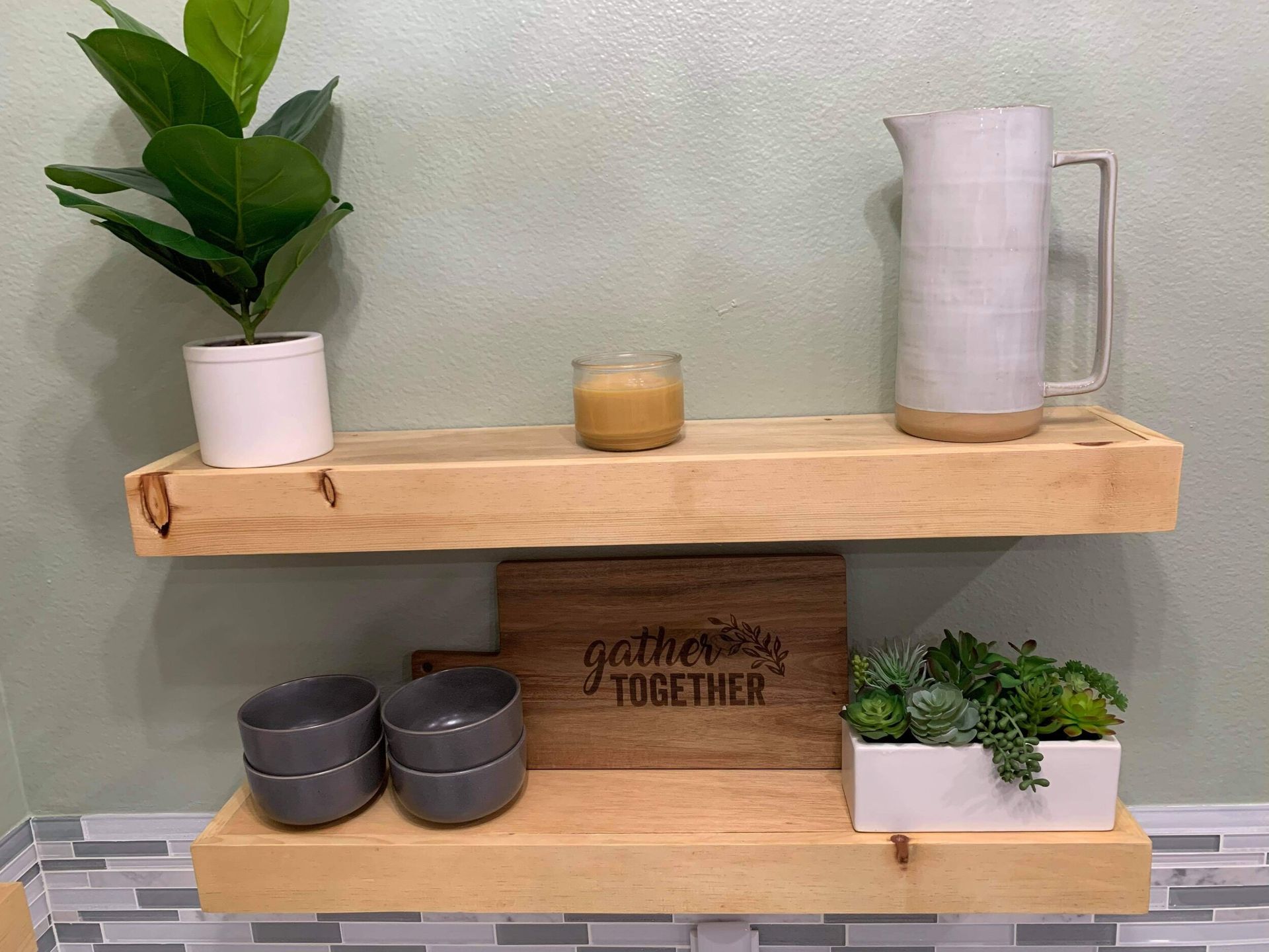 Two wooden shelves with decor: plant, candle, pitcher, bowls, and a cutting board that says, 