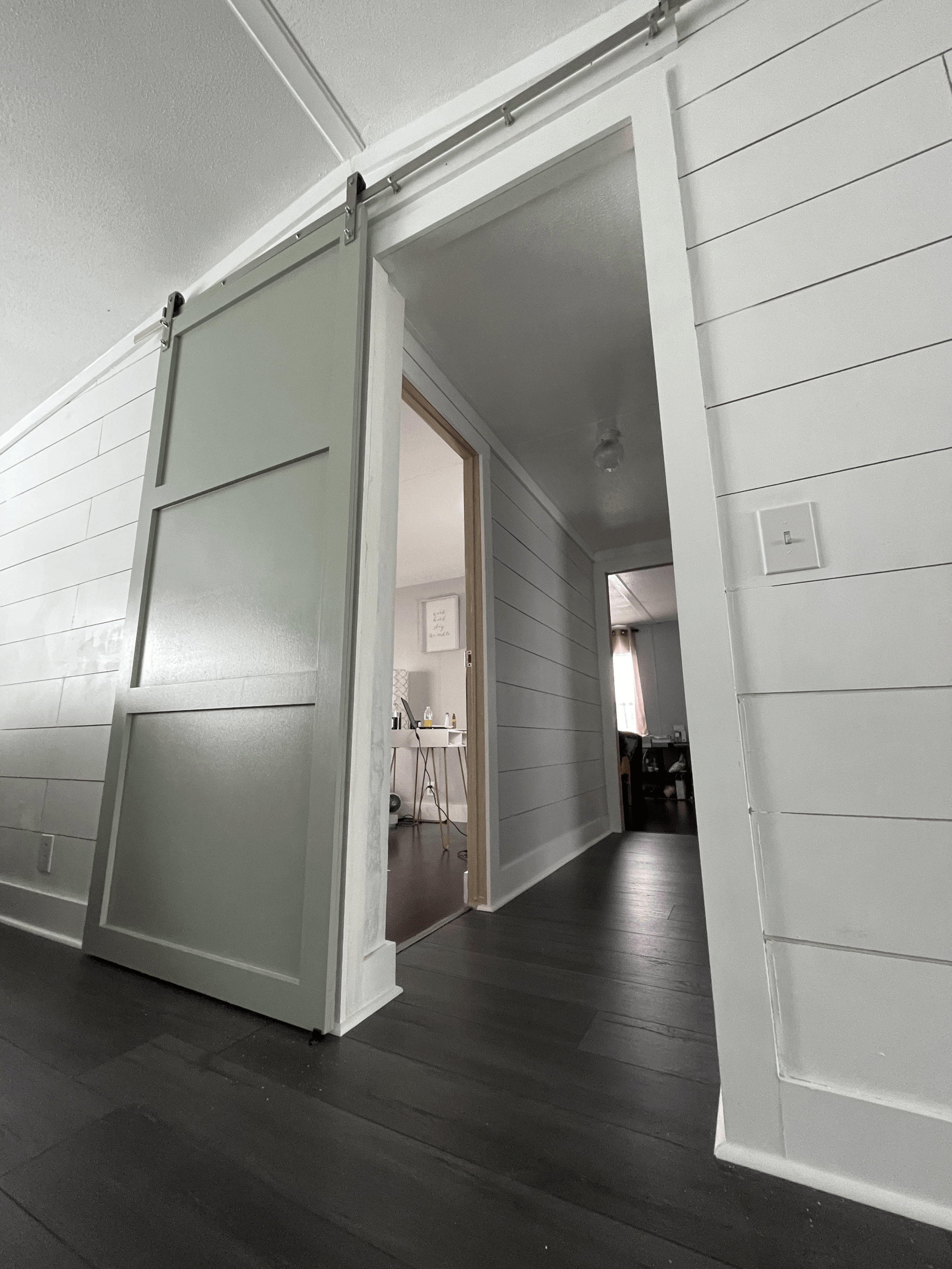 Ajar gray sliding barn door leads to a hallway with white paneled walls and dark flooring.