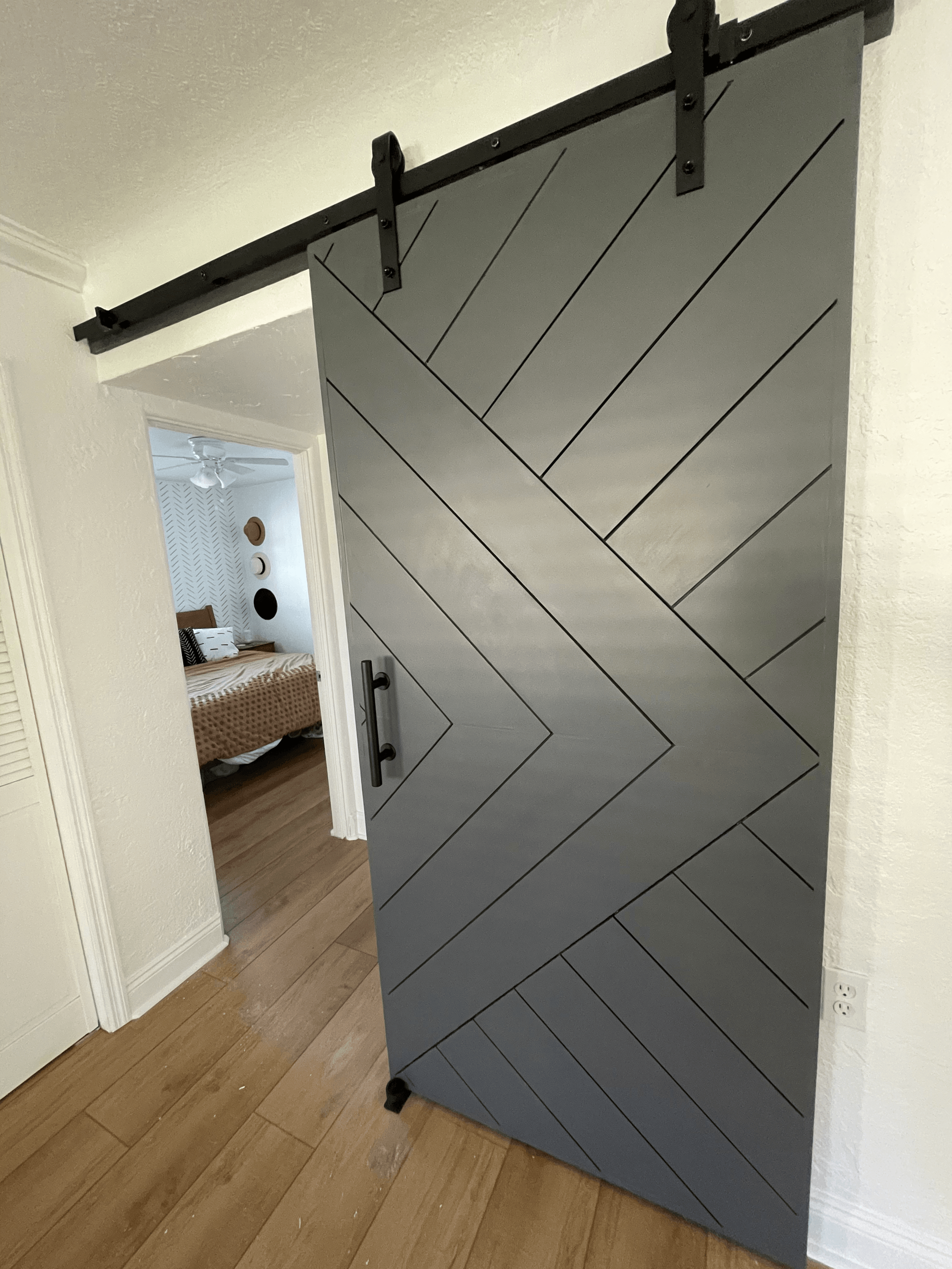 Gray herringbone pattern barn door open into a bedroom, black hardware, mounted on a rail.