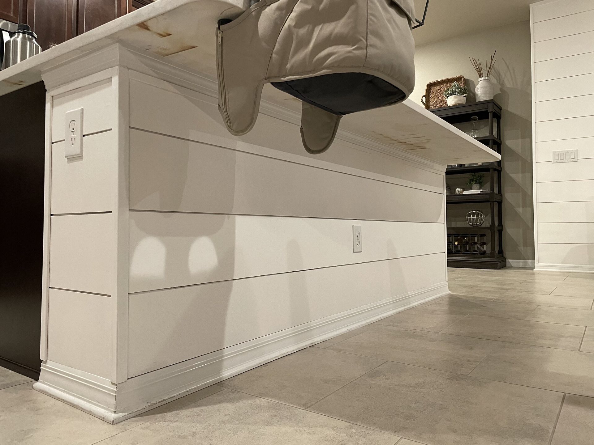 White kitchen island with horizontal plank siding and outlet, oven mitts hanging.