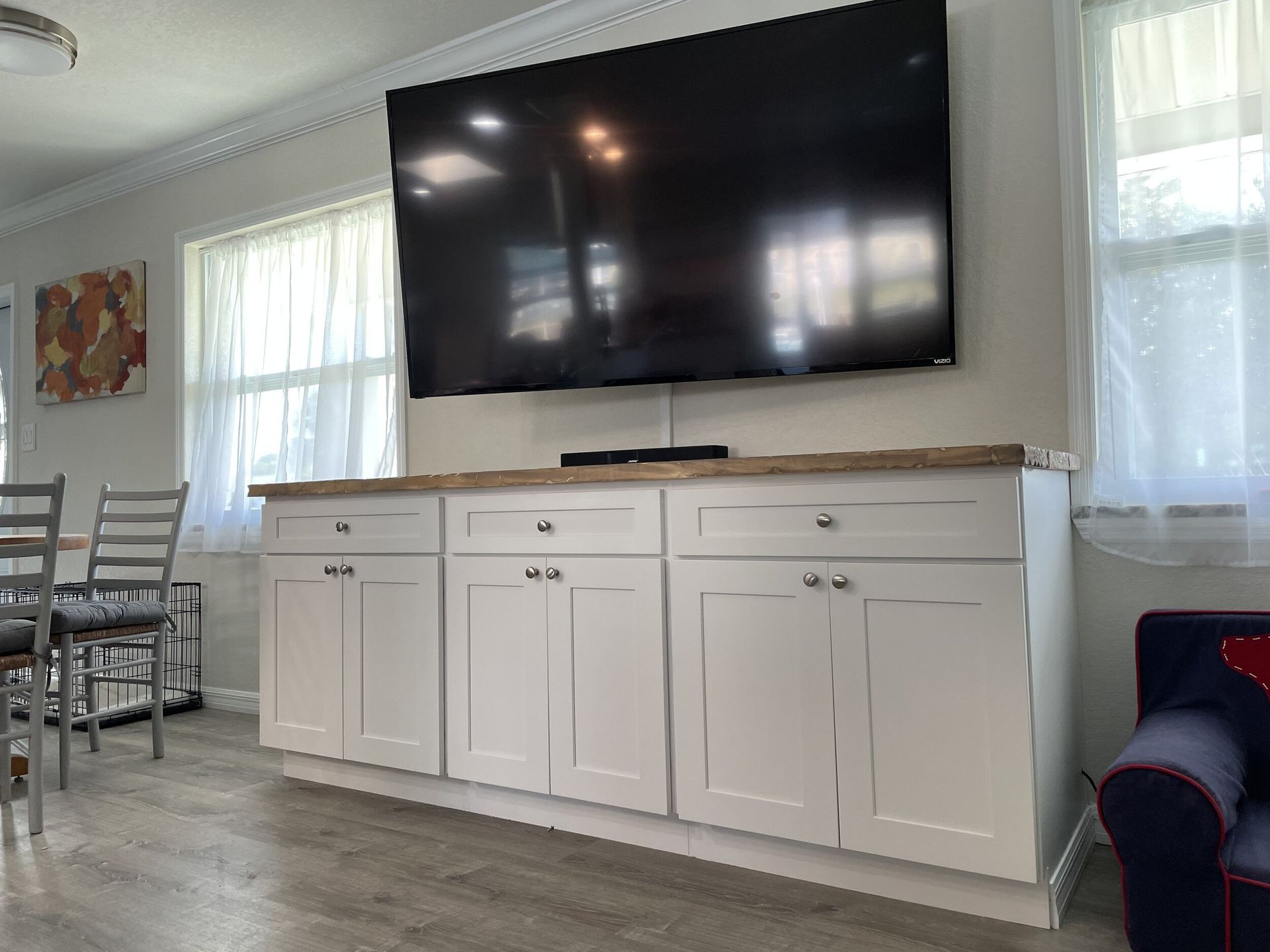 White cabinets with TV above. Windows on either side. Blue couch on the right. Dining area to the left.