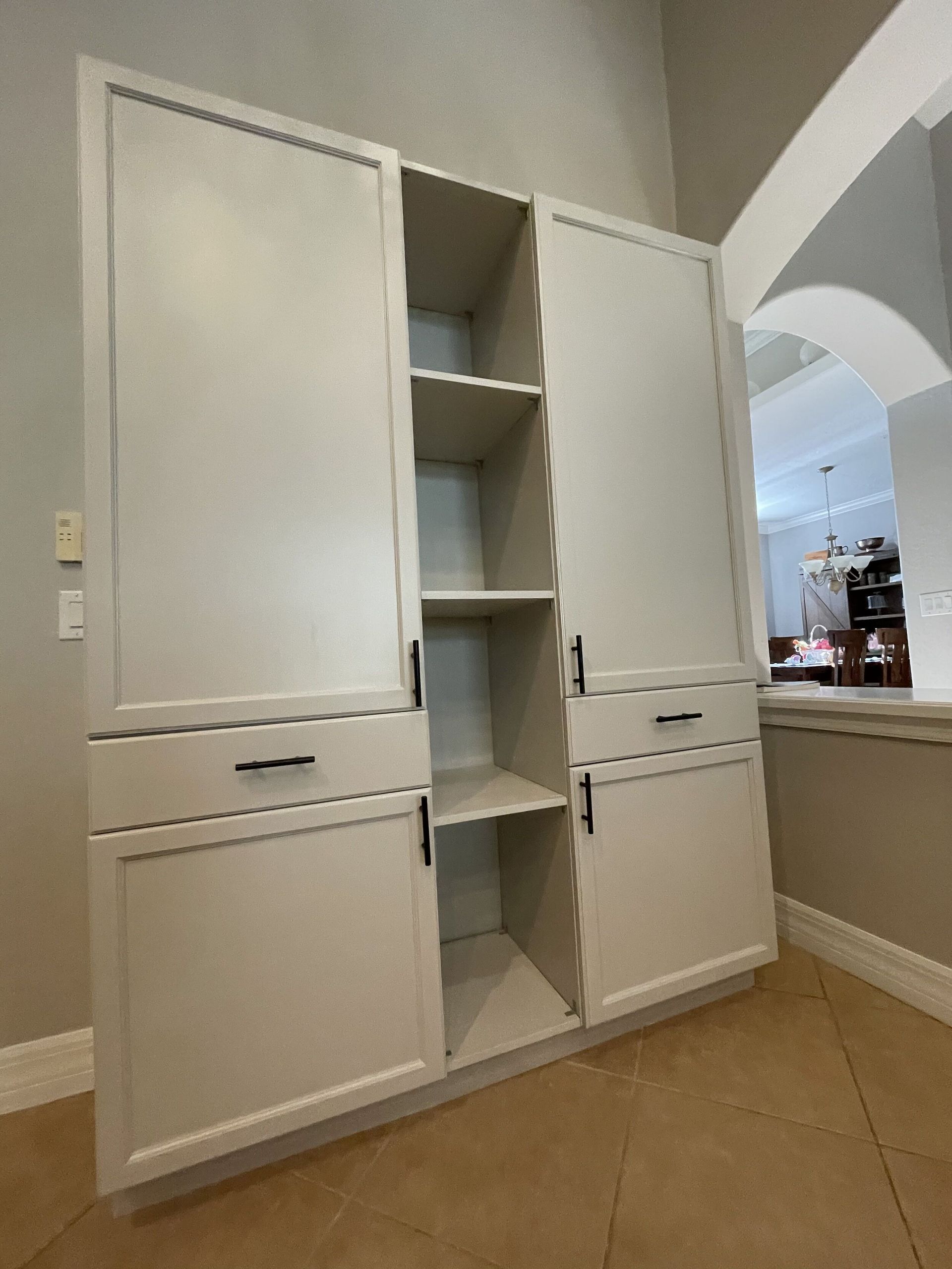 Light-colored kitchen pantry with two tall doors and open shelves. Dark handles. Arched doorway visible.