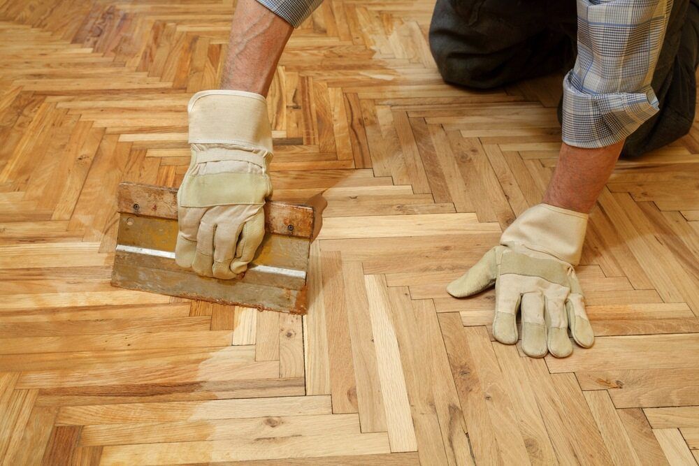 A Man is Painting a Wooden Floor With a Spatula — Shane Callinan Floor Sanding In Forster - Tuncurry, NSW