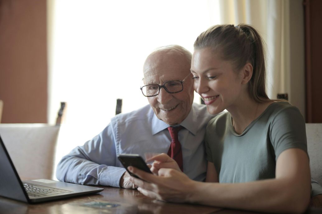 An elderly man and a young woman are sitting at a table looking at a cell phone.