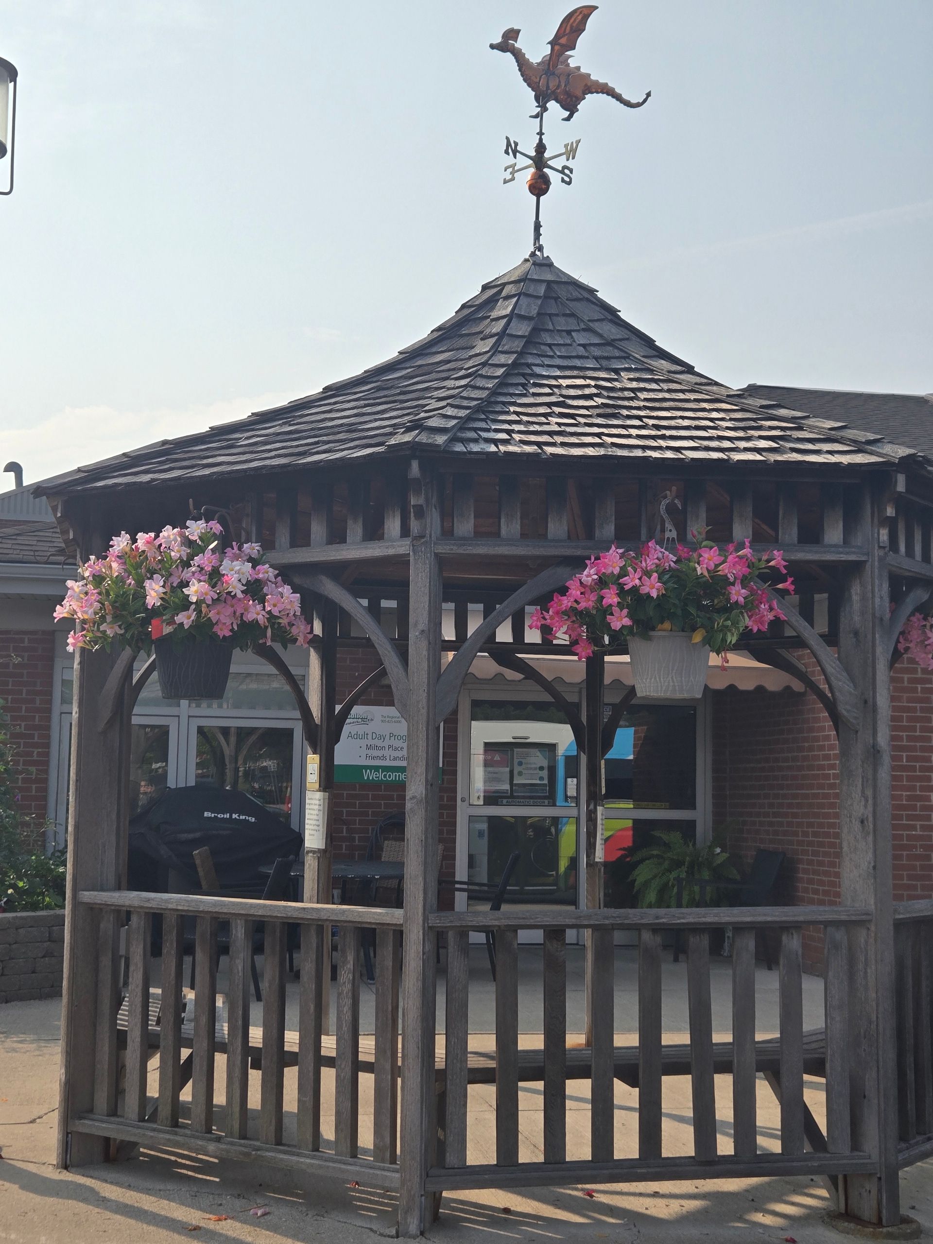 Wooden gazebo with a dragon weathervane, flowers, and a brick building.