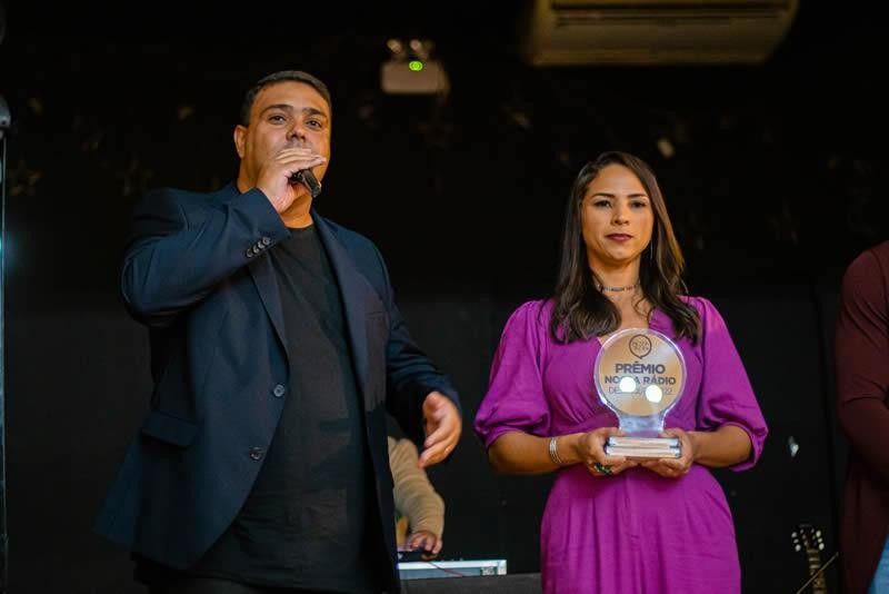 Homem fala ao microfone, mulher segura prêmio. Eles estão no palco em um ambiente escuro.