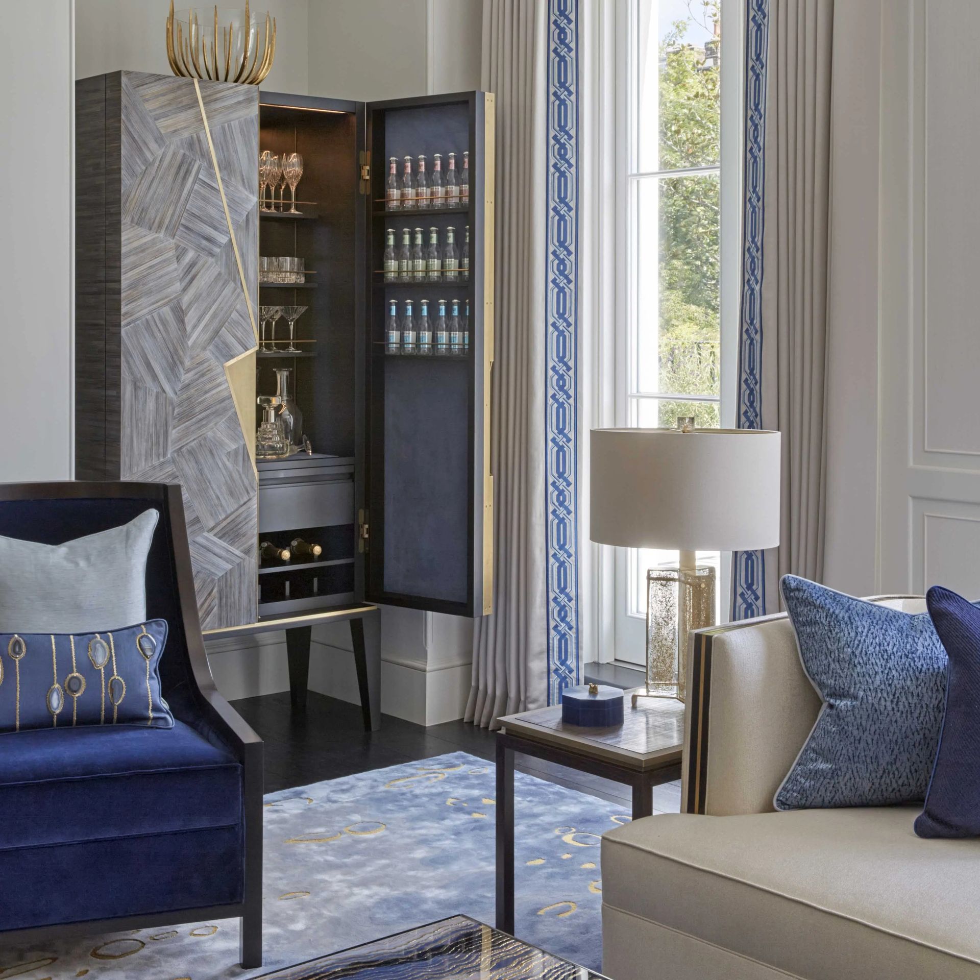 Living room with blue accents, featuring a bar cabinet, velvet chair, and patterned rug.
