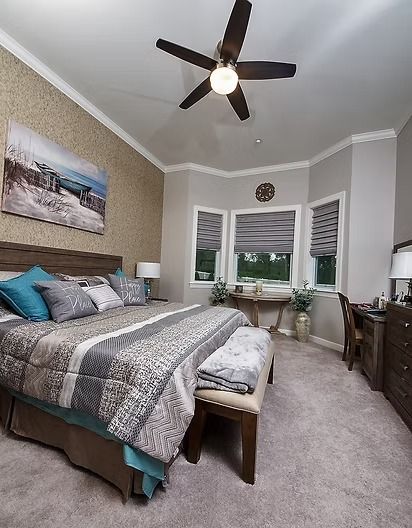 Bedroom with neutral tones, bed, artwork, desk, windows, and ceiling fan.