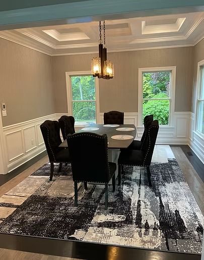 Dining room with a table, six black chairs, and a patterned rug. Windows and a chandelier are visible.