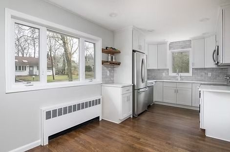 Modern kitchen with white cabinets, stainless steel appliances, and dark wood floors. Large window overlooks a yard.