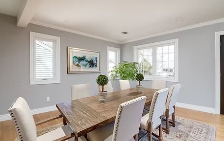 Dining room with wooden table, white chairs, and large windows. Gray walls and a rug.