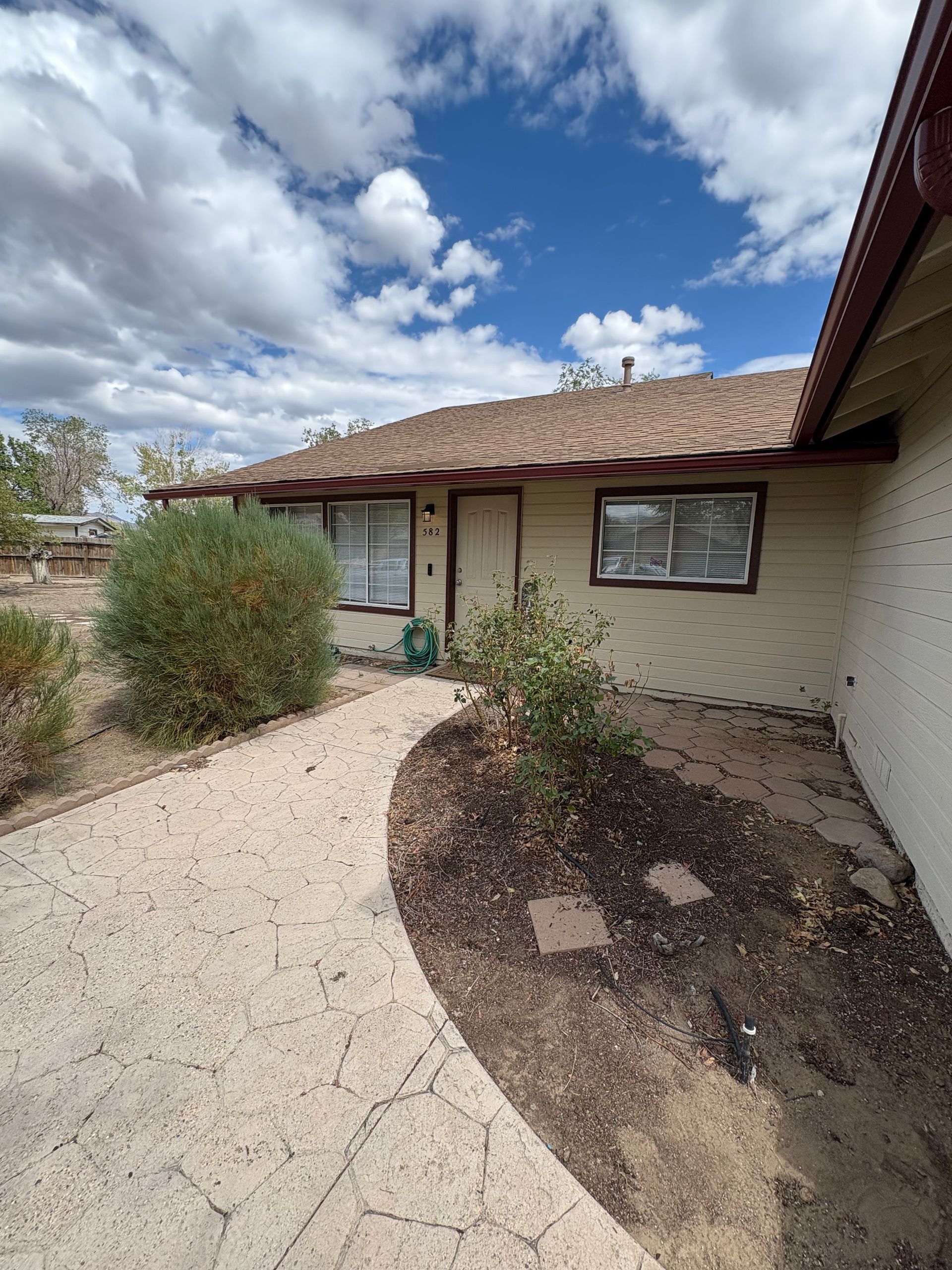 Tan house with brown roof, walkway, and landscaping against a blue sky with clouds.