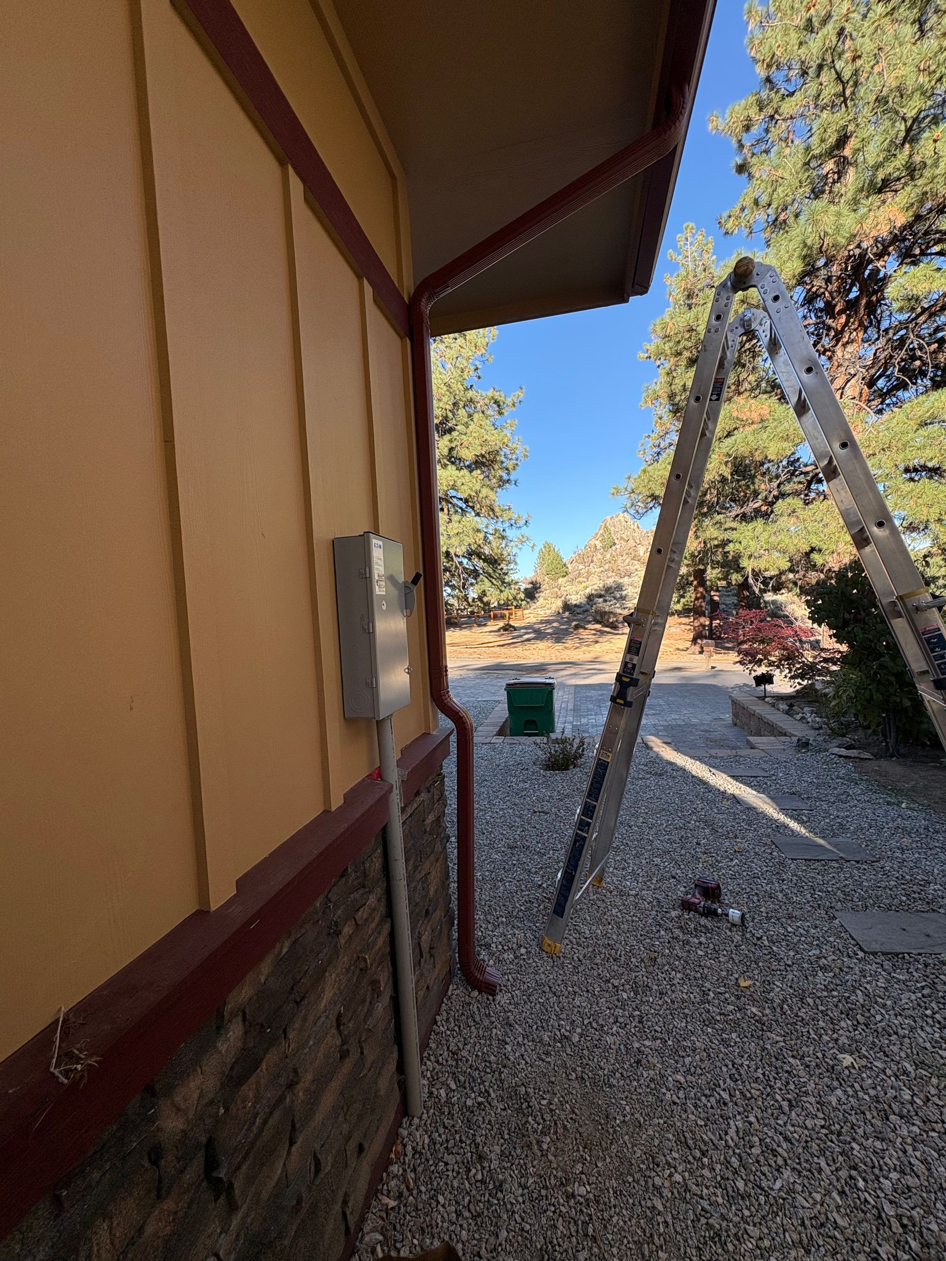 Yellow house exterior with ladder, electrical box, and gravel ground. Blue sky with trees in background.
