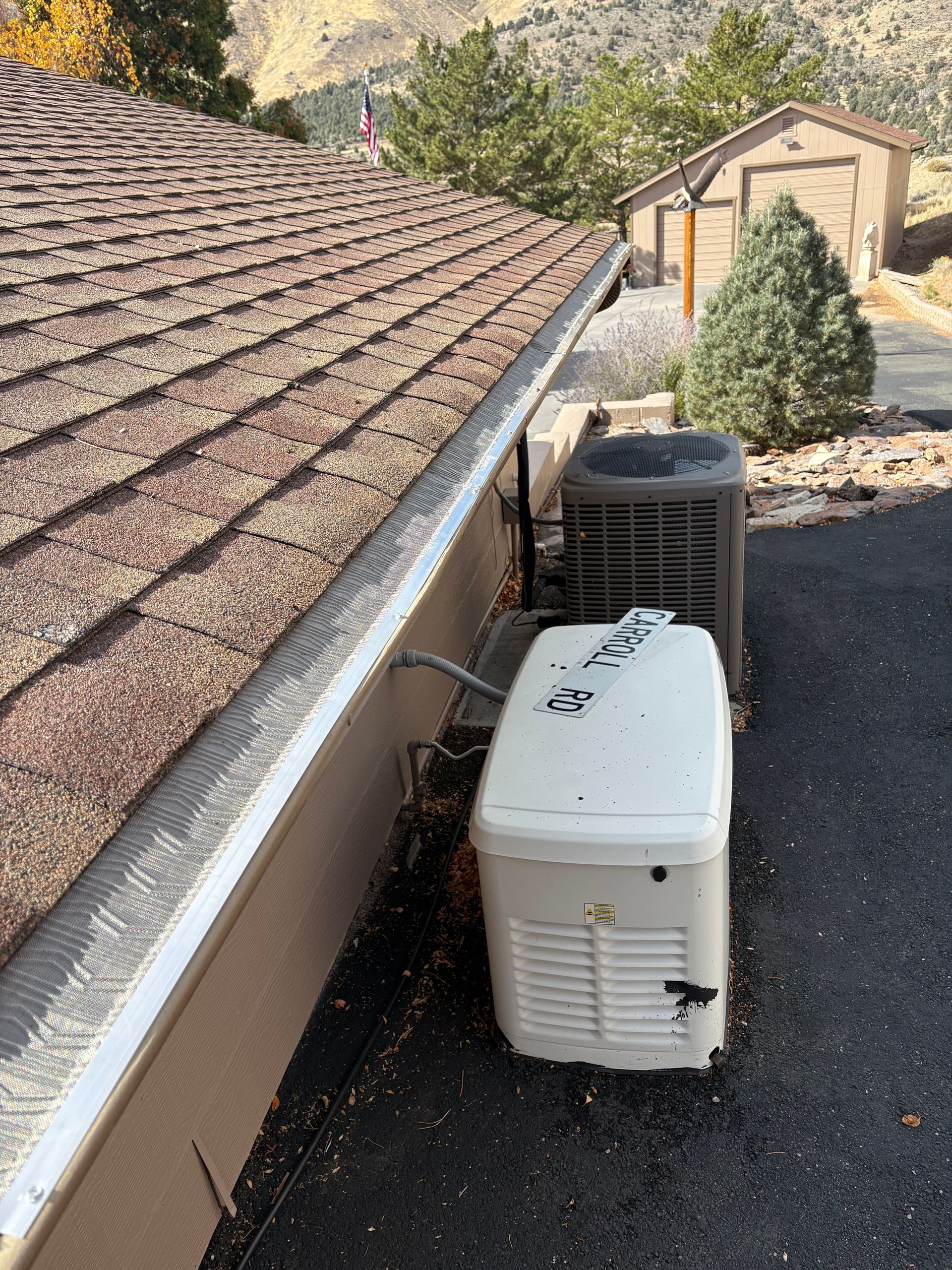 Asphalt roof with gutter guard, generator, and air conditioner on blacktop. Building in background.